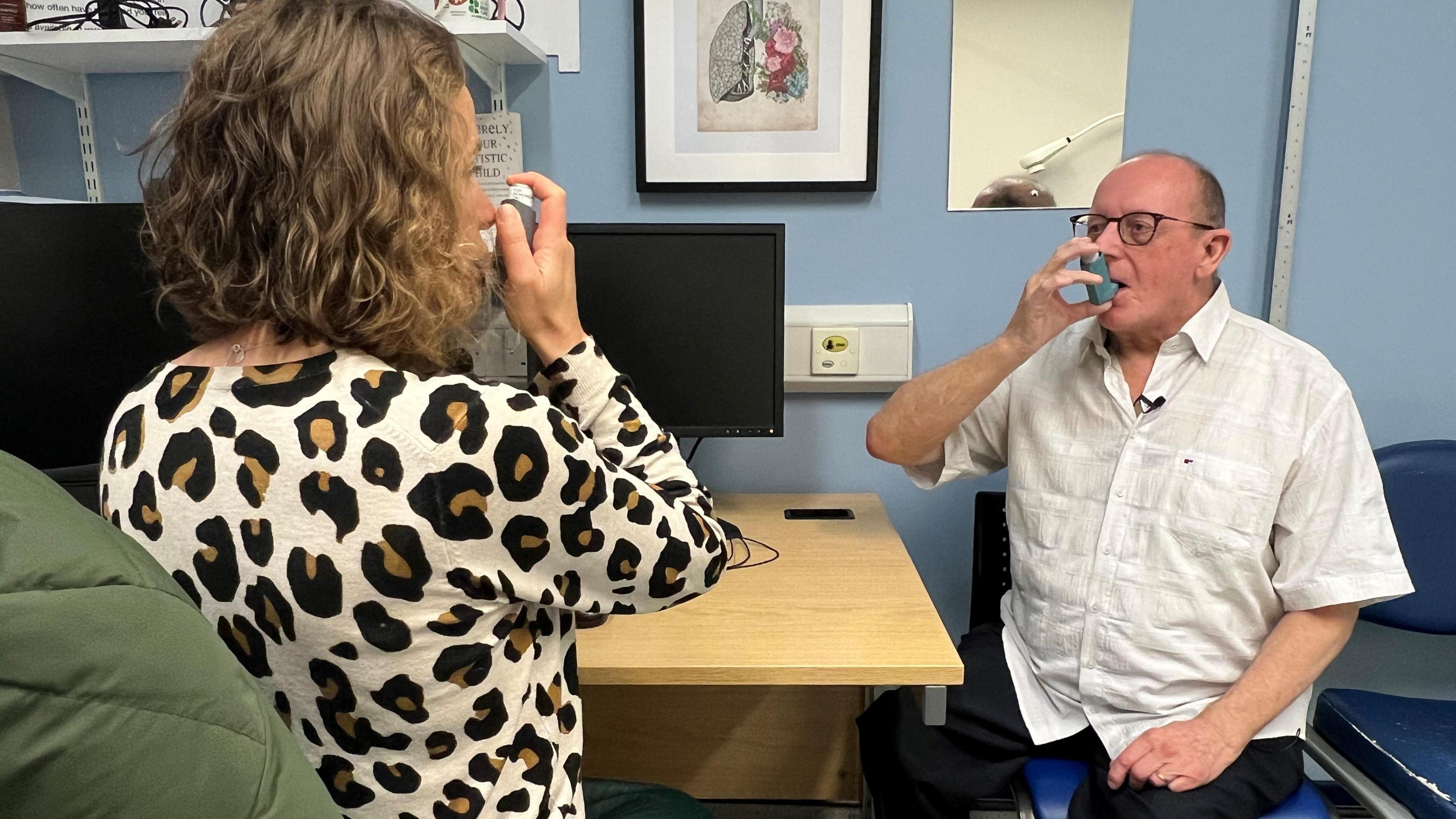 A bespectacled gentleman wearing a white shirt uses an inhaler during a GP appointment. A woman on the other side of the desk demonstrates the inhaler.