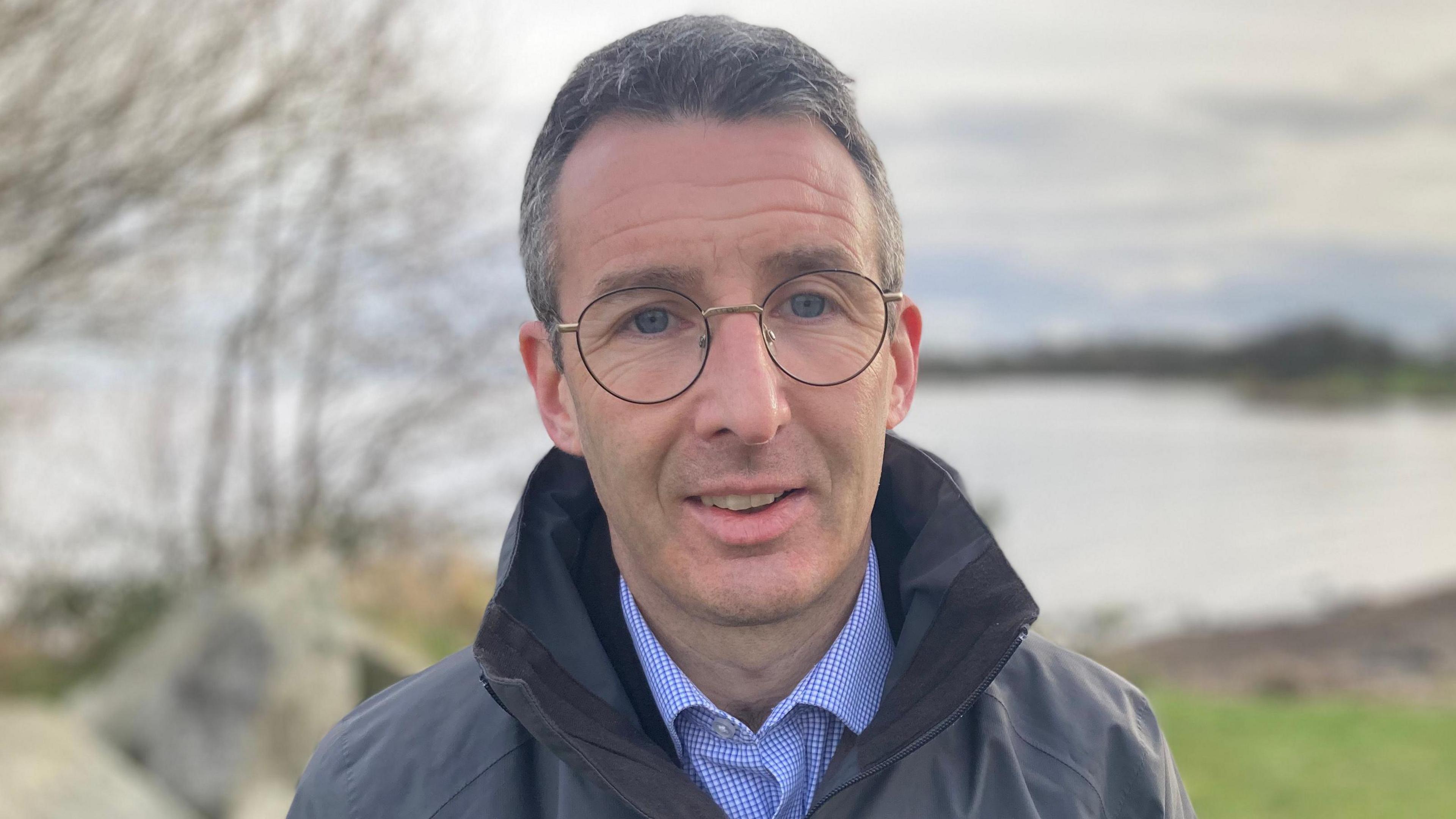 Andrew Mui, who has short grey-black hair and is wearing round glasses. He's standing in front of a lake. He's wearing a blue and white small-checked shirt and a grey coat raincoat.