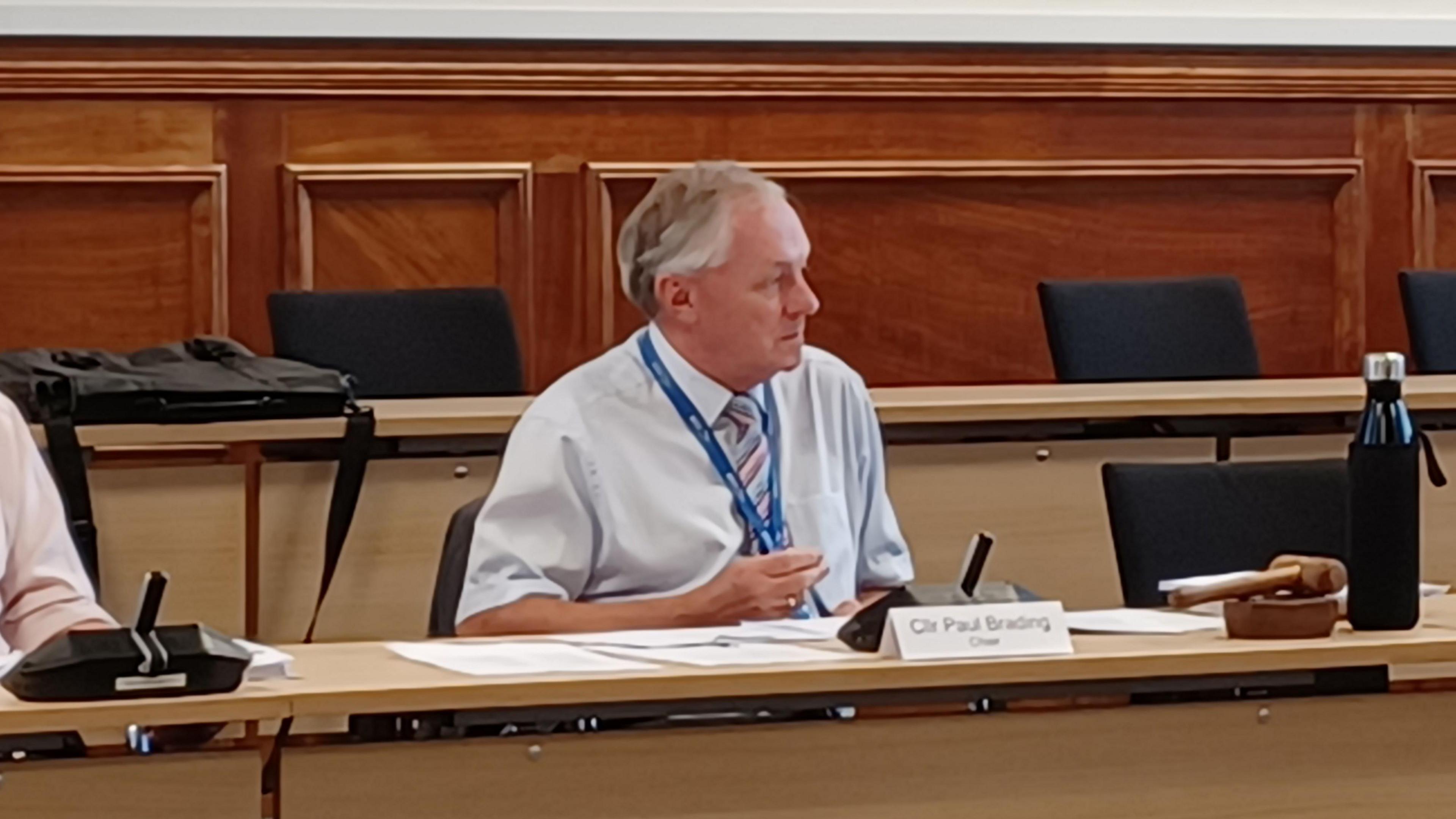 Conservative education committee chair Paul Brading in a white/light blue shirt with sleeves rolled up, and a stripy tie, sitting behind a meeting room bench