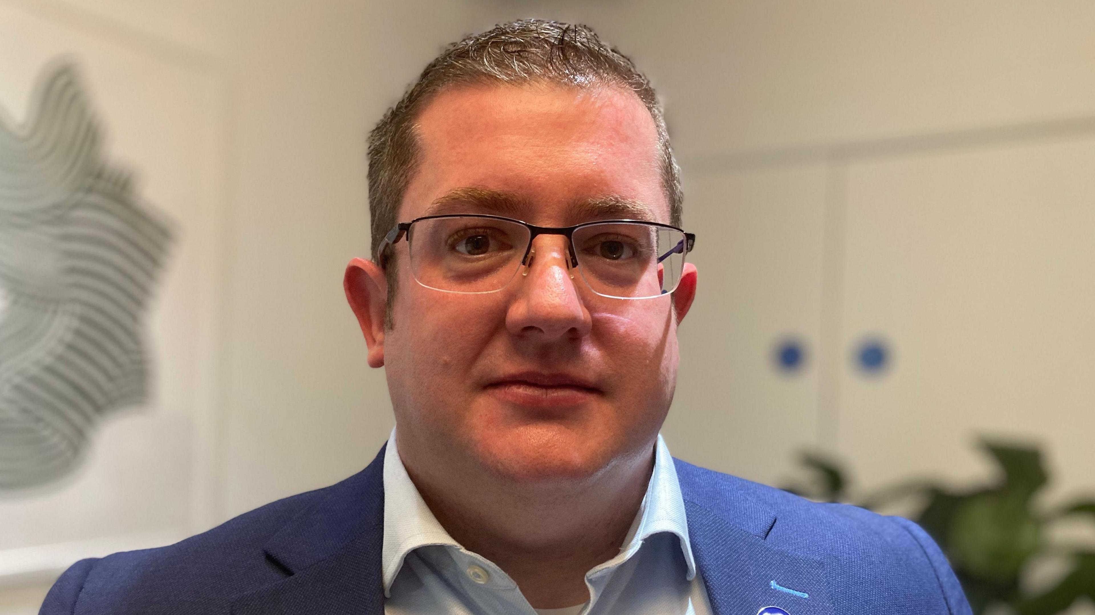 White background, focus on David. He is wearing square glasses, he has short grey/black hair and is wearing a light blue shirt and navy suit jacket. 