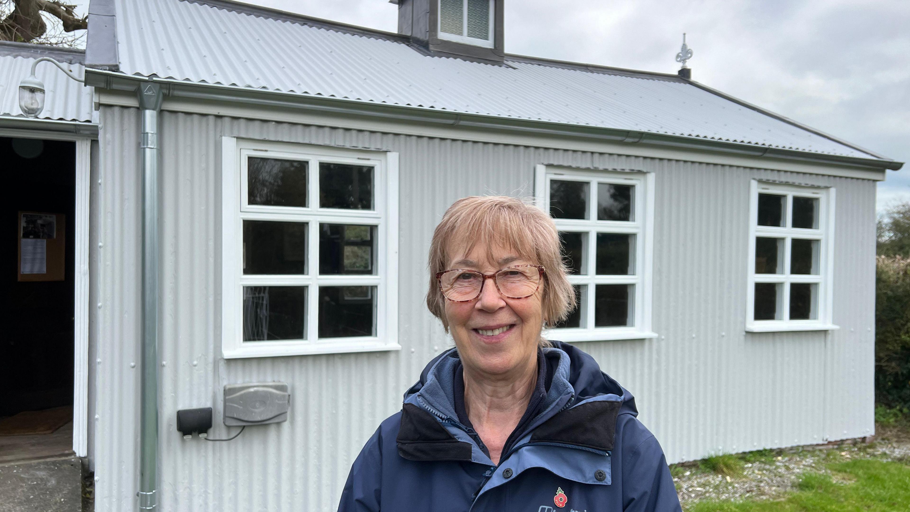 Maureen Phillips is standing outside the little tin church. She is wearing a blue jacket and has short hair. The church has a tin exterior and three windows on the side.