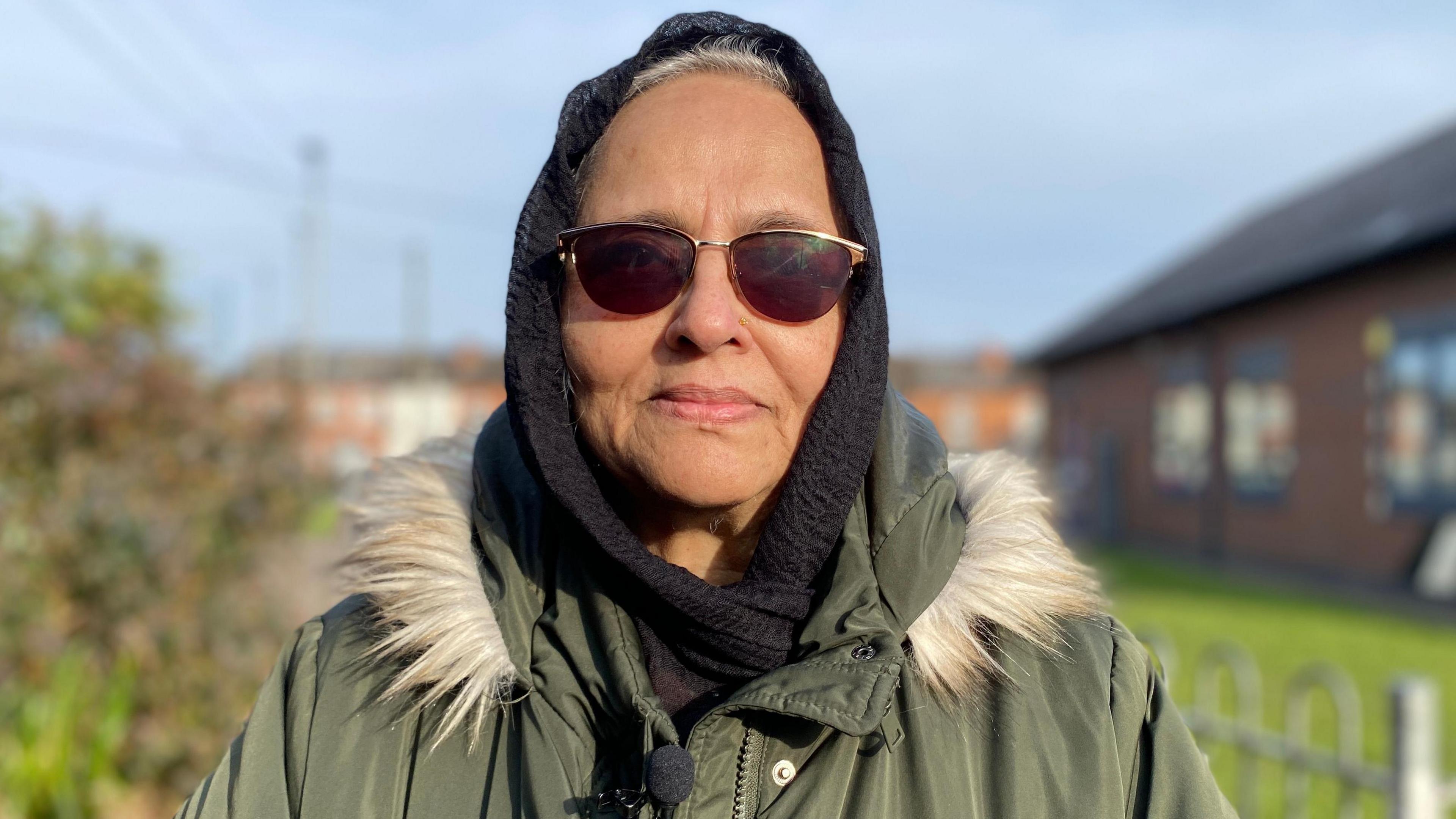 Dureshahwar Shaikh is wearing a green coat with a furry hood. She has sunglasses covering her eyes and a black hijab on her head. 