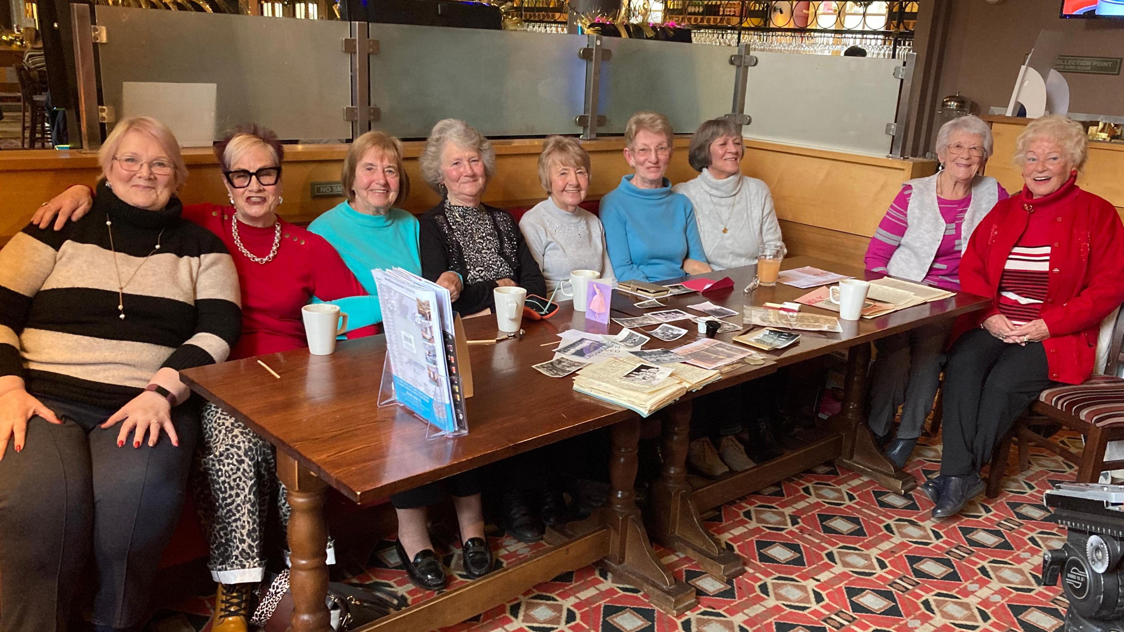 Nine women, aged in their 70s and 80s, smile for the camera as they sit around a large table in a pub in Hull. Photos, old schools books and other pieces of memorabilia are laid out on the table in front of them.