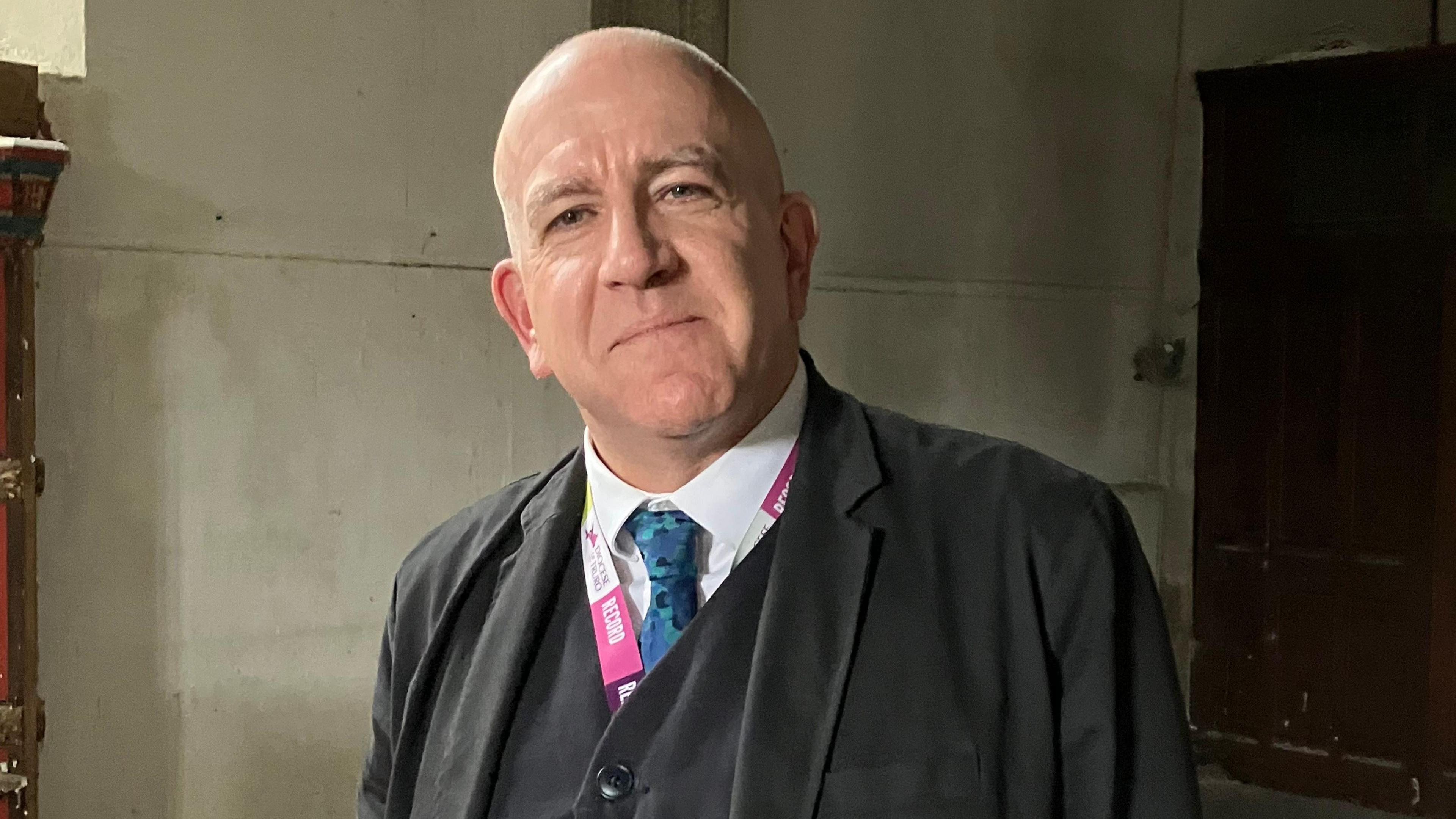 Simon Cade, who is smiling at the camera while wearing a black suit and vest with a blue tie and white shirt. He has a partly visible pink lanyard around his neck. In the background is a white stone wall.