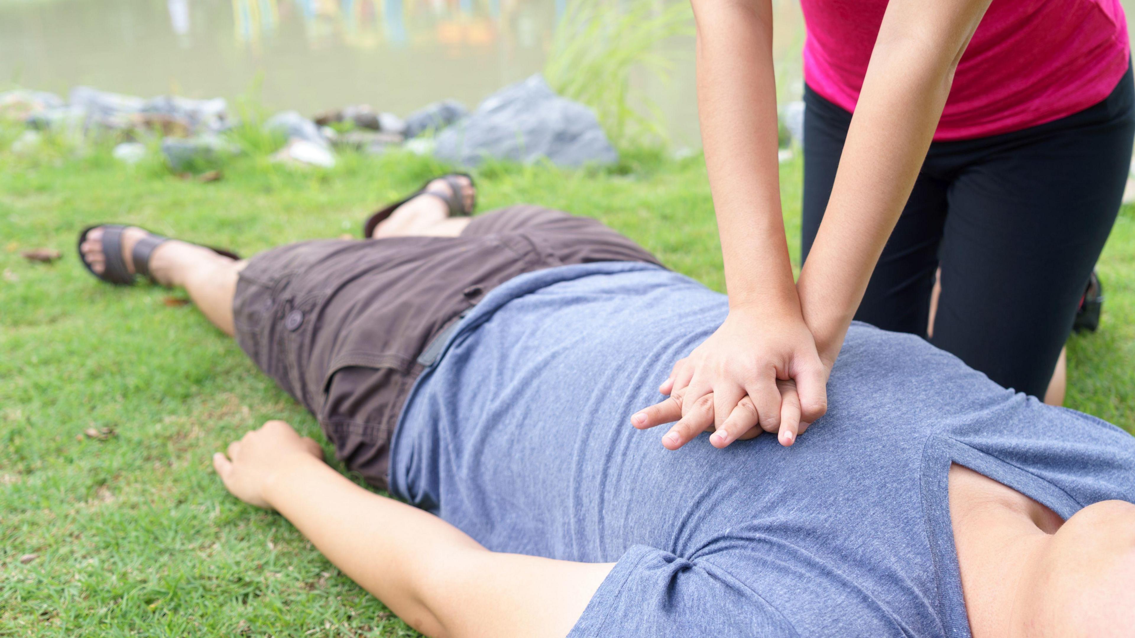 Woman giving cardiopulmonary resuscitation (CPR) to a man at a public park. The man is lying on the grass wearing a blue T-shirt and grey shorts. A woman is kneeling over him wearing a pink top and black leggings. She has her hands with fingers linked over his chest. 