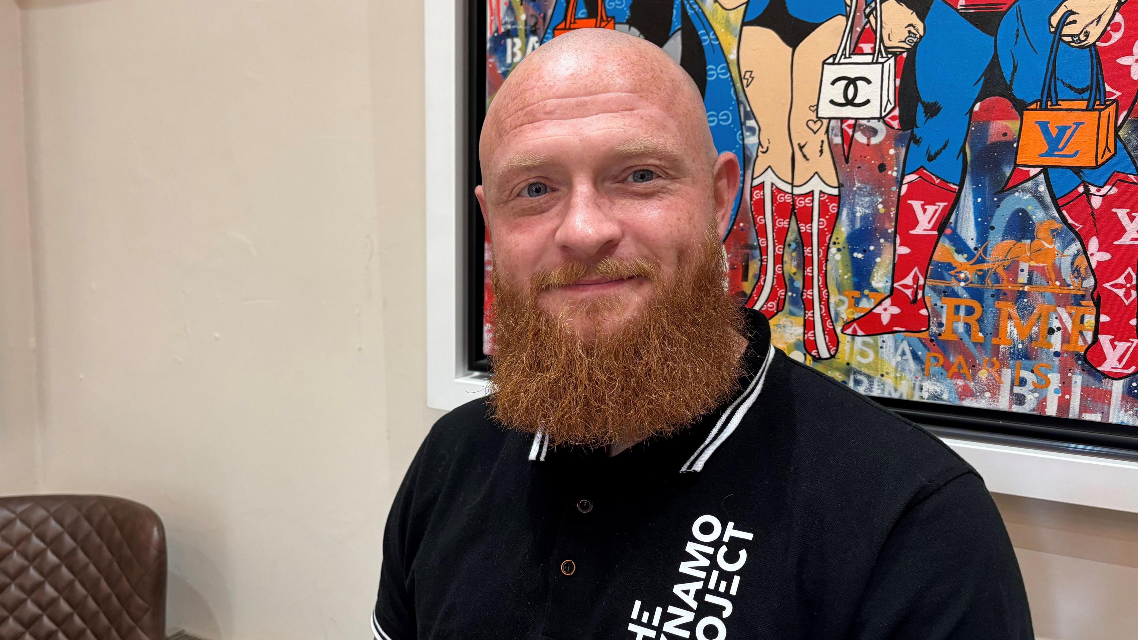 A man who is bald, wearing a black T-shirt and has a long ginger beard. He is smiling at the camera and sitting in front of a brightly coloured picture that is hanging on the wall.