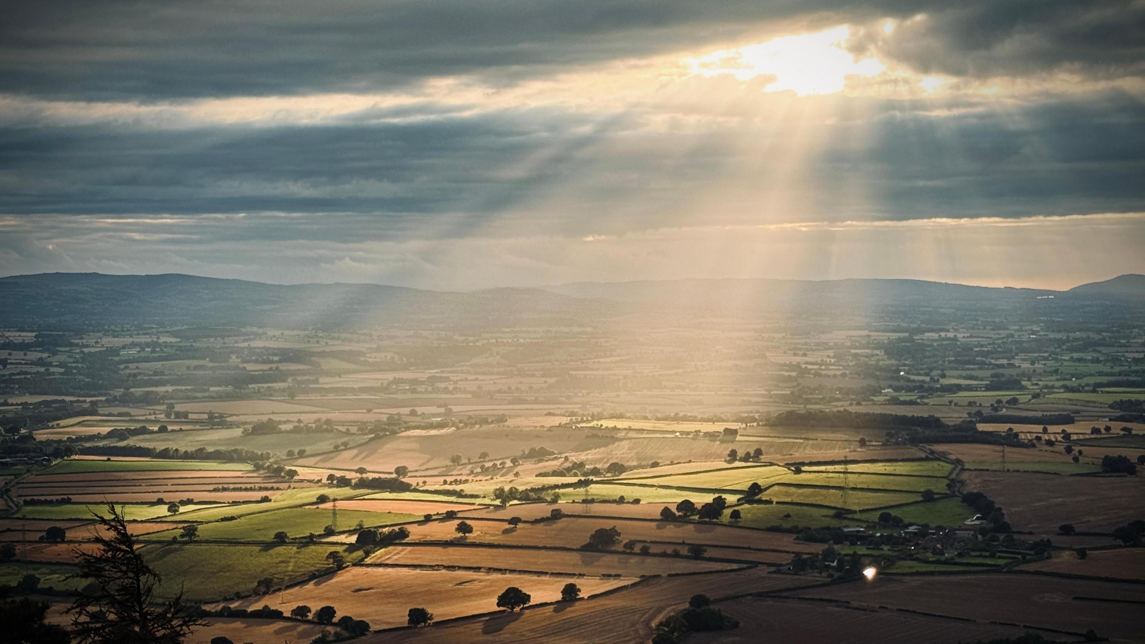 Sunbeams break through grey clouds and splay out over fields of green and golden crops with hills in the background.