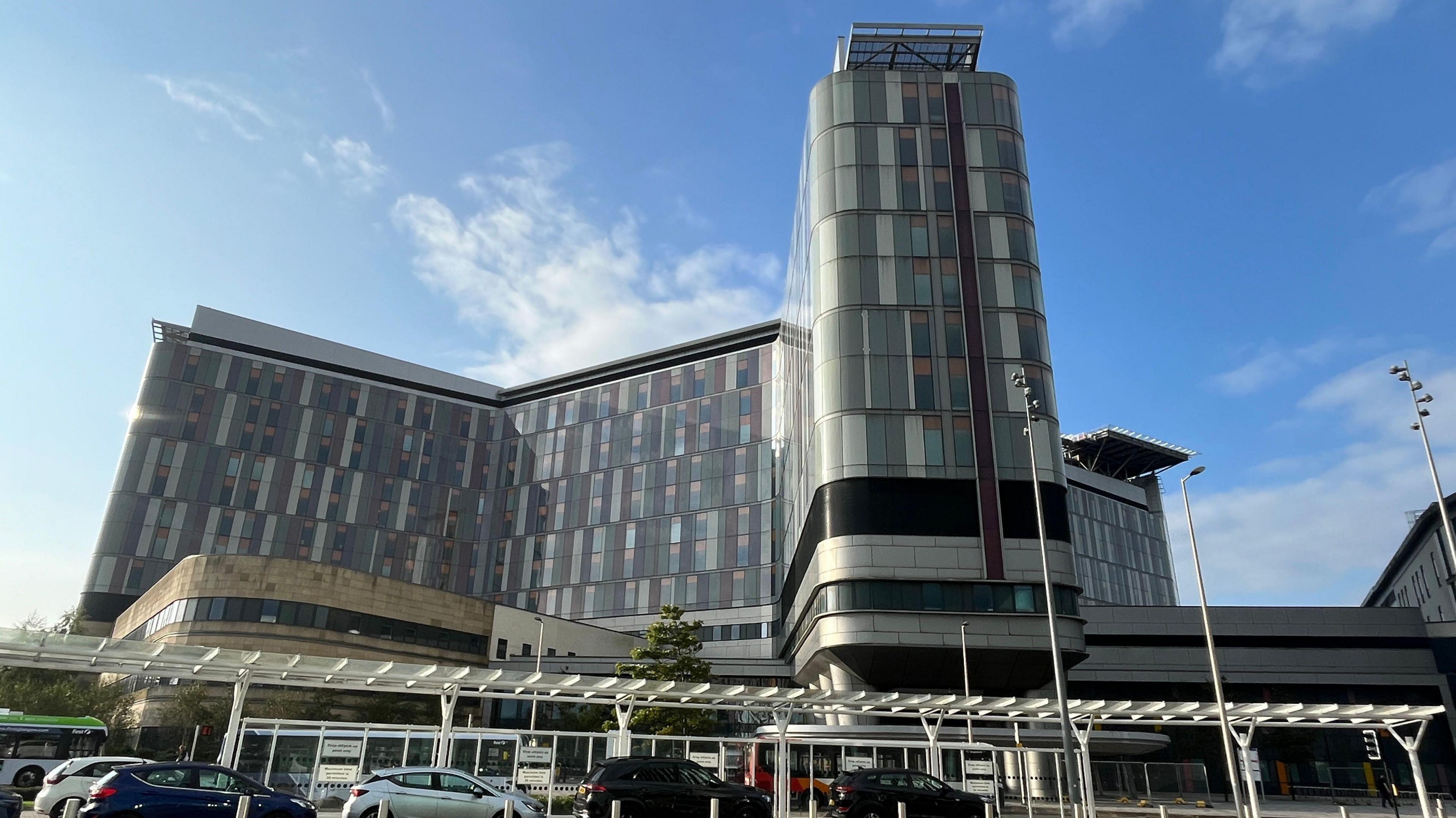 The outside of the Queen Elizabeth University Hospital on a sunny day. The picture is looking upwards at the main hospital building, which is in front of a blue sky with a few white clouds overhead.