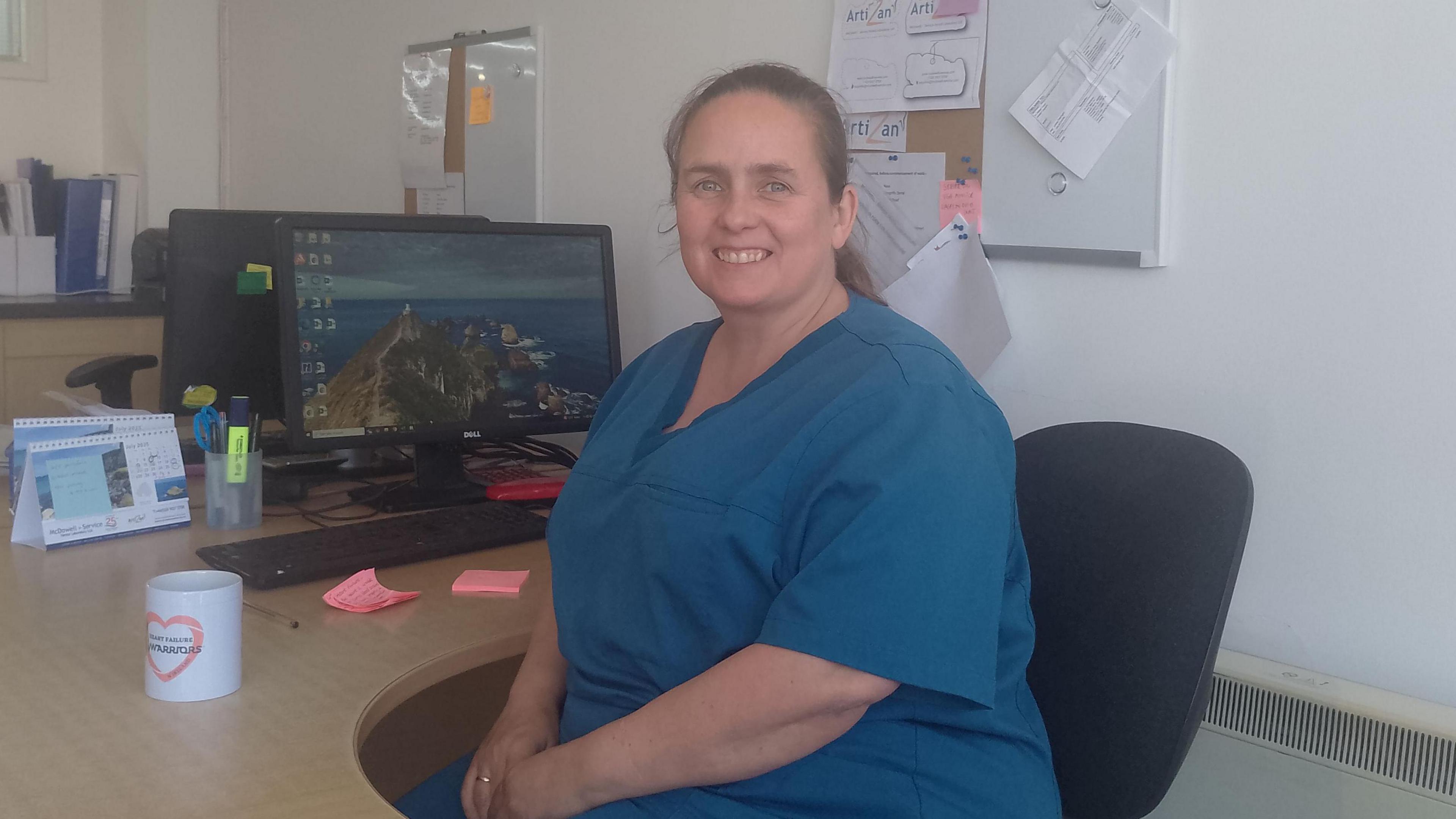 Nicole McKelvie has long brown hair tied back and is wearing blue scrubs and sitting at a desk with a computer monitor