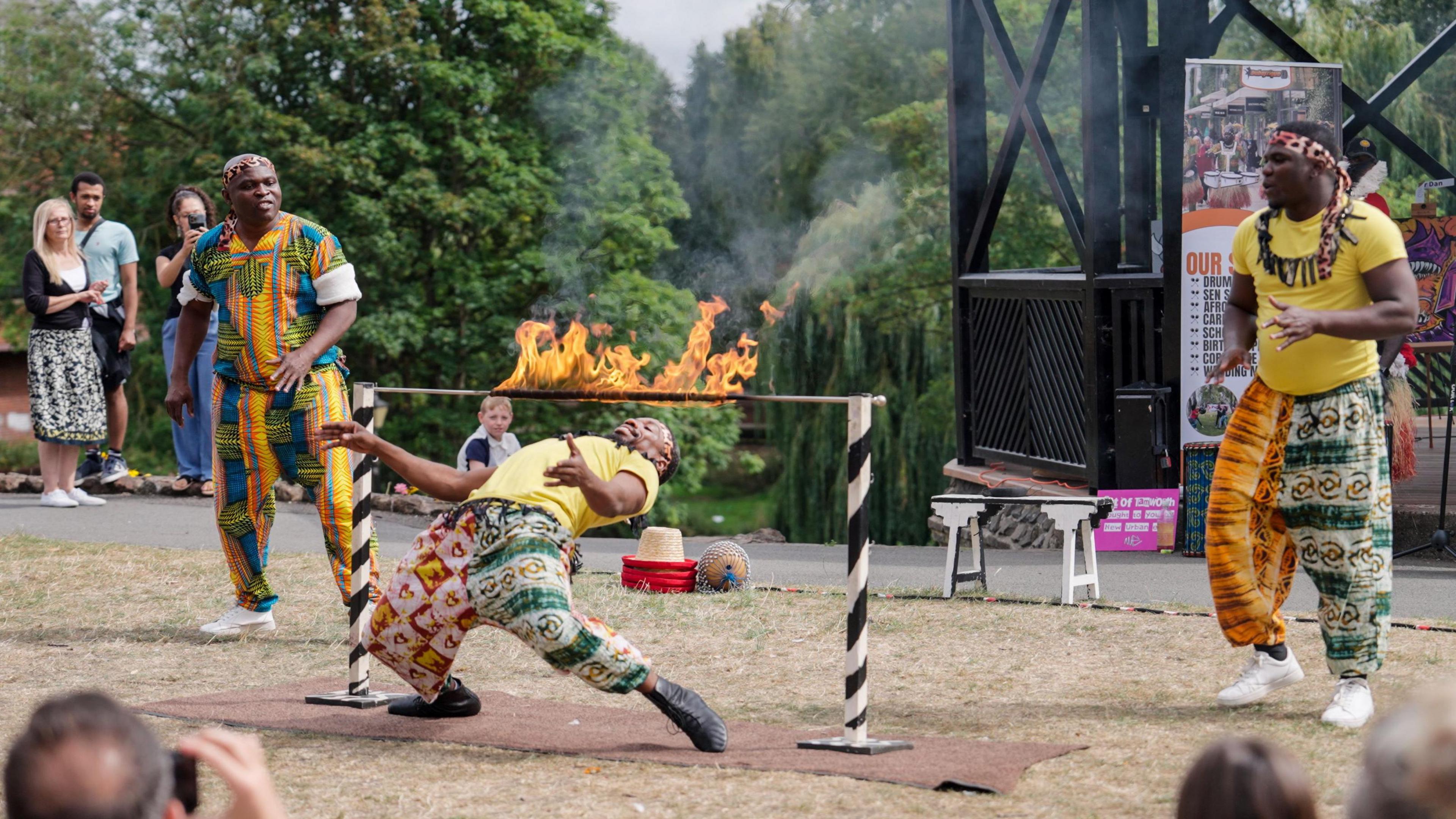 A man does a limbo dance under a pole which is one fire. He is watched by two other performers and several onlookers in the background. 