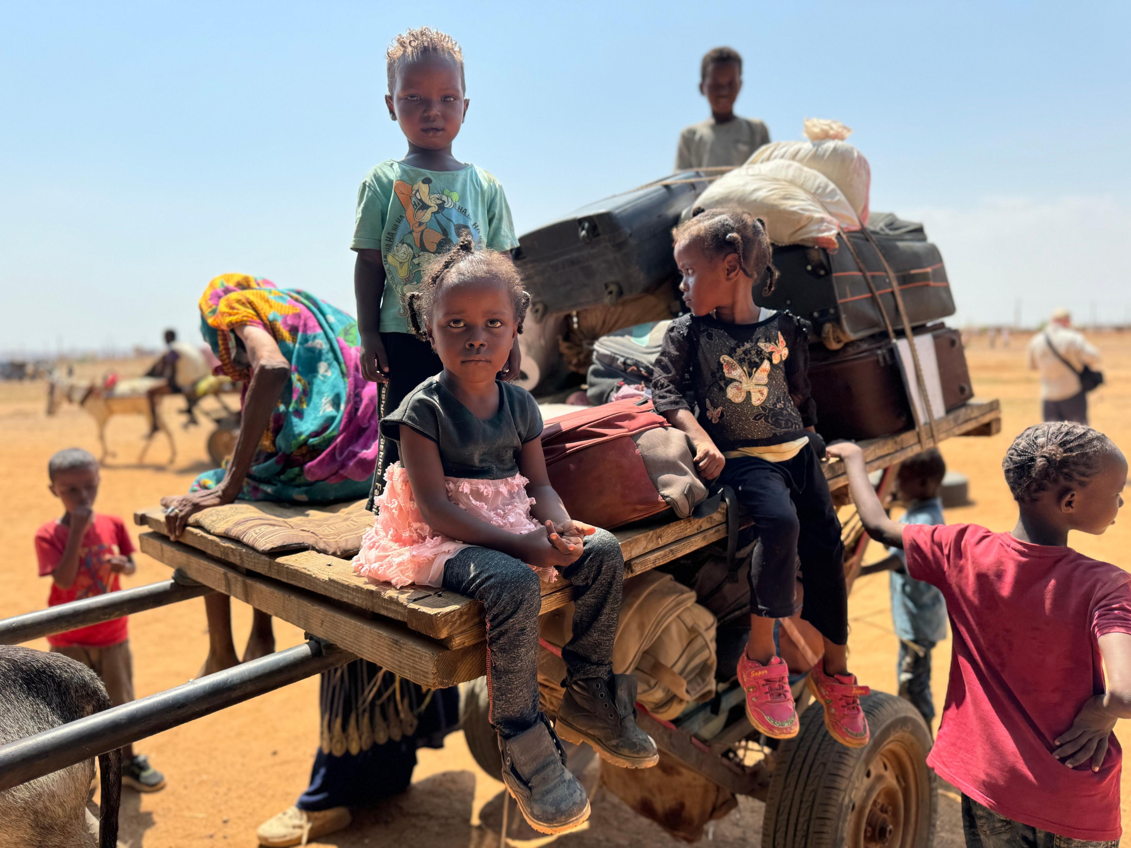 Young children sitting on a wooden wagon which is also laden with suitcases in a hot and dusty environment