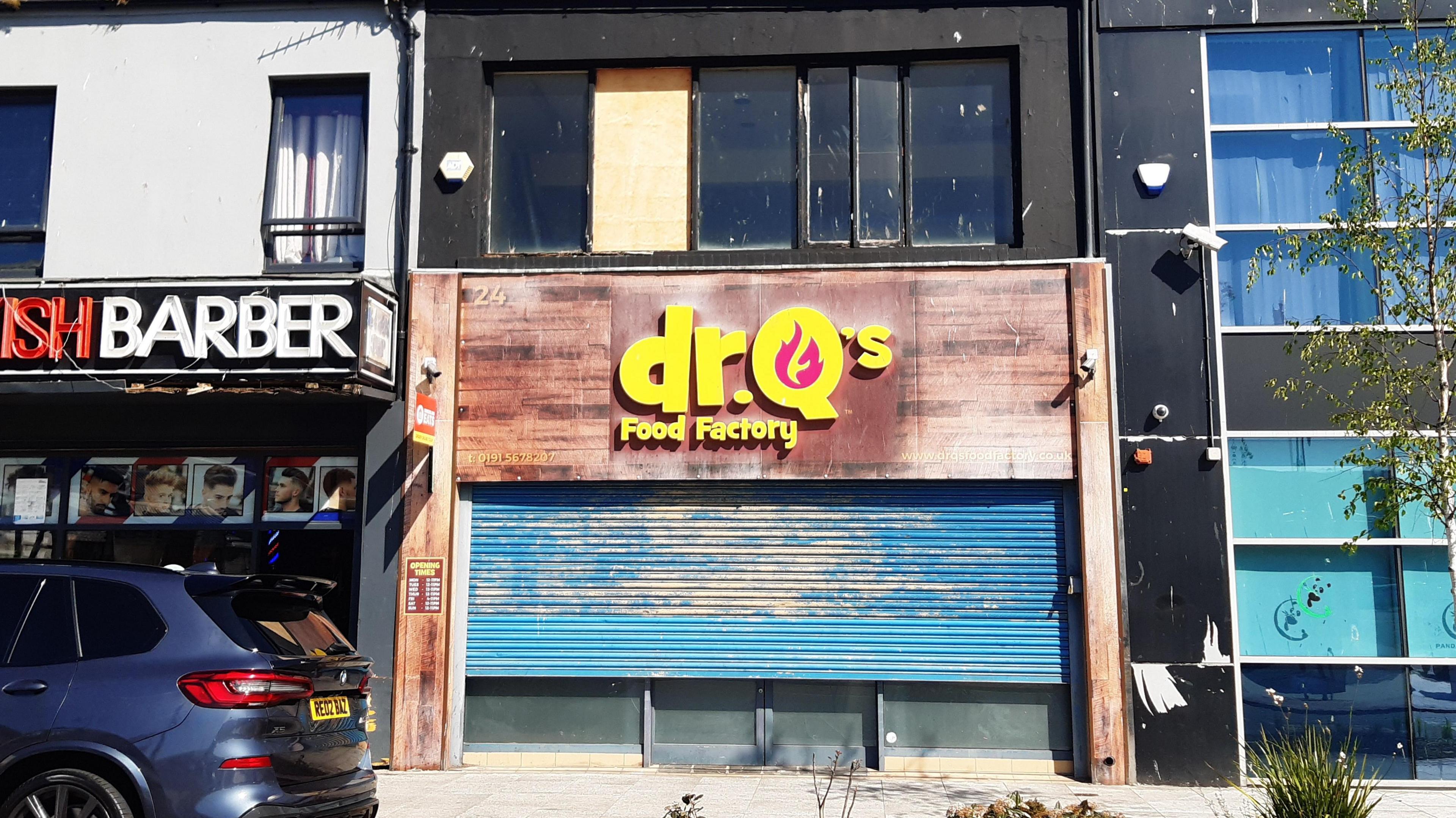 Dr Q's Food Factory which has a brown shop front with blue shutters which have not been pulled all the way down. A glass-fronted building can be seen to the right and a Turkish barber shop to the left. The flats would have sat above Dr Q's.