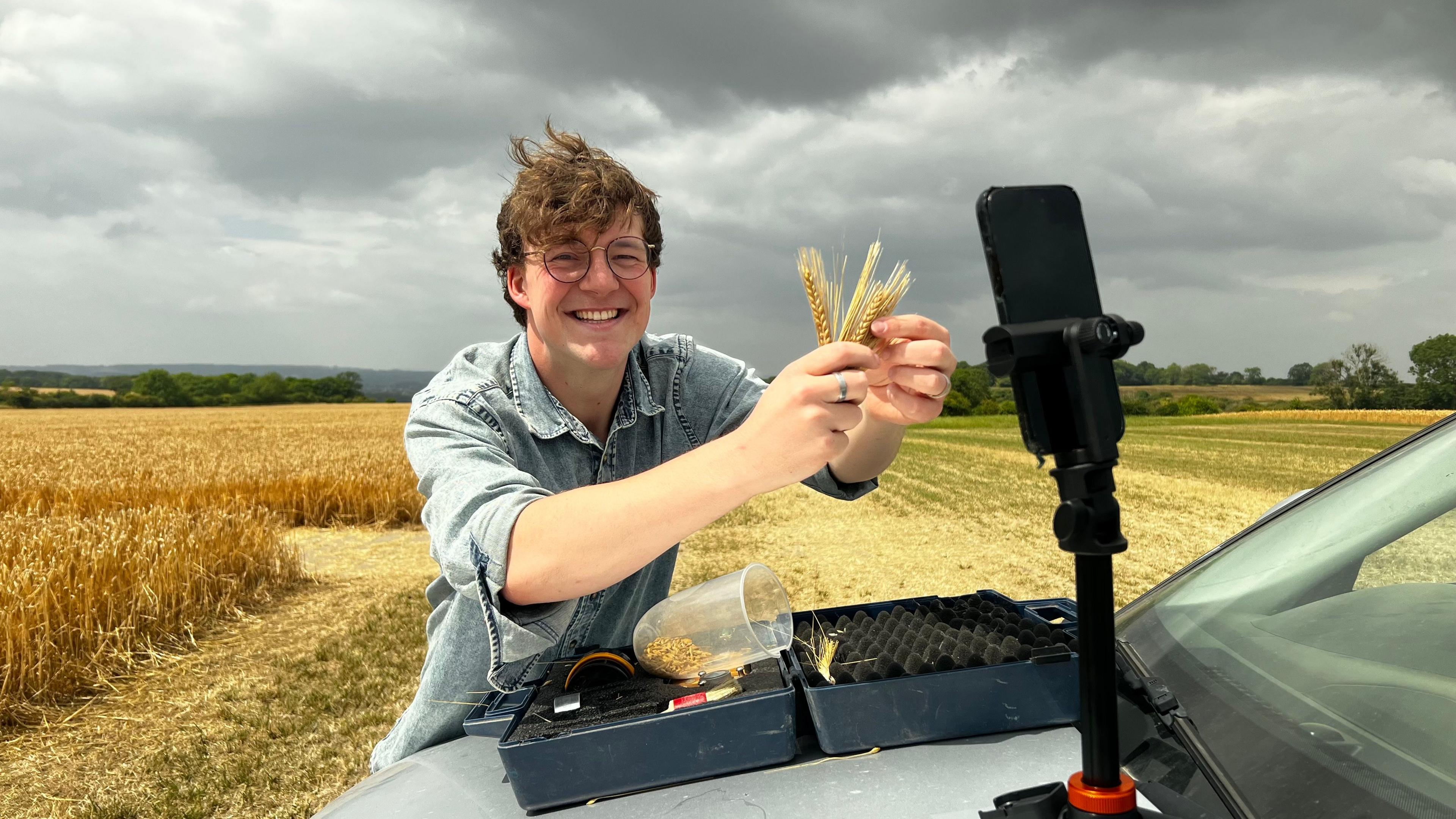 Mike Wilkins smiles at the camera as he holds up fresh ears of barley to his phone camera. The phone is fastened to a tripod which is balanced on the boot of his silver truck. The car is parked in a field with a cloudy sky behind.