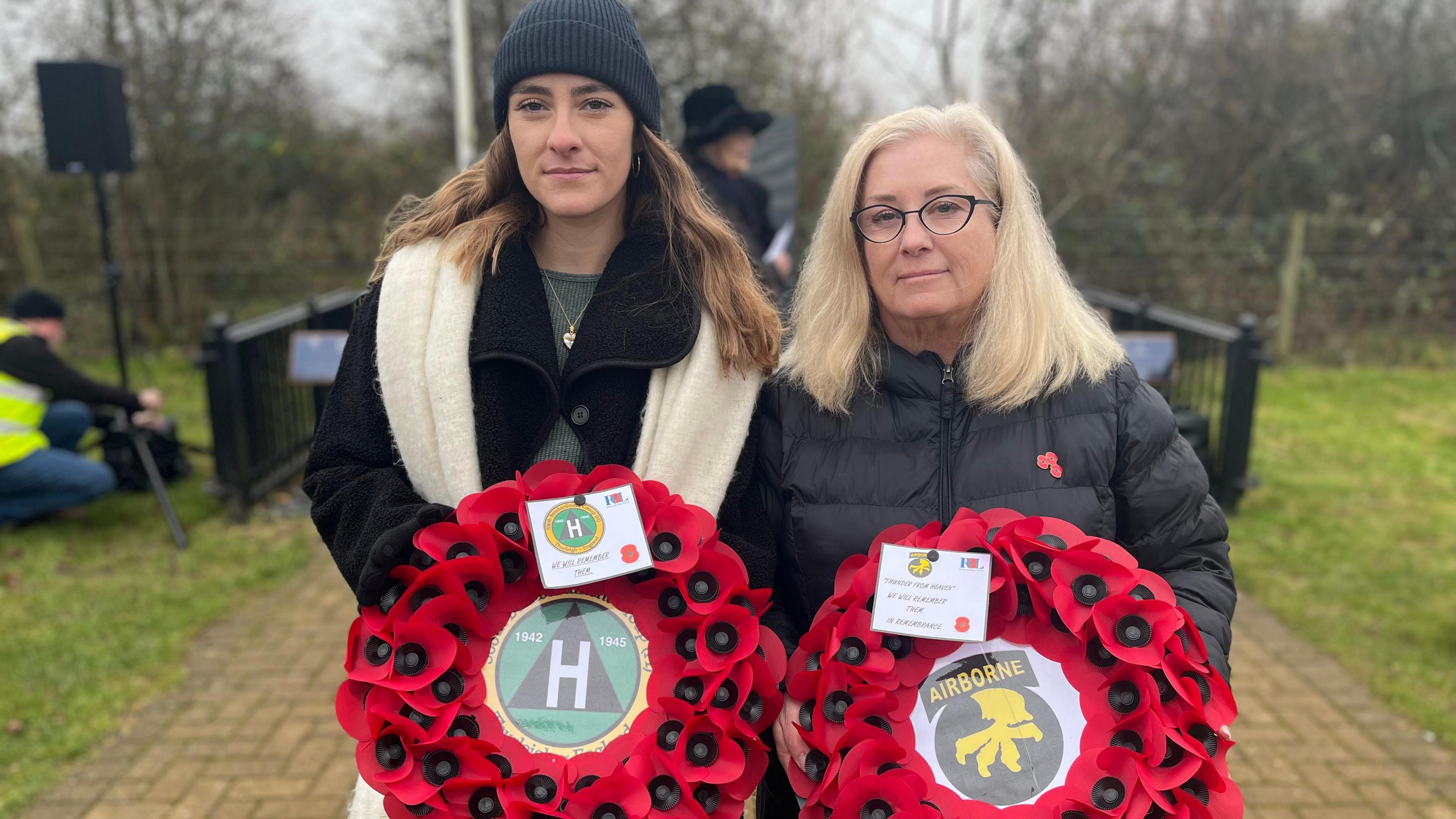 Two blonde women wearing black coats holding up poppy wreaths and looking straight at the camera. There's grass and trees behind them.