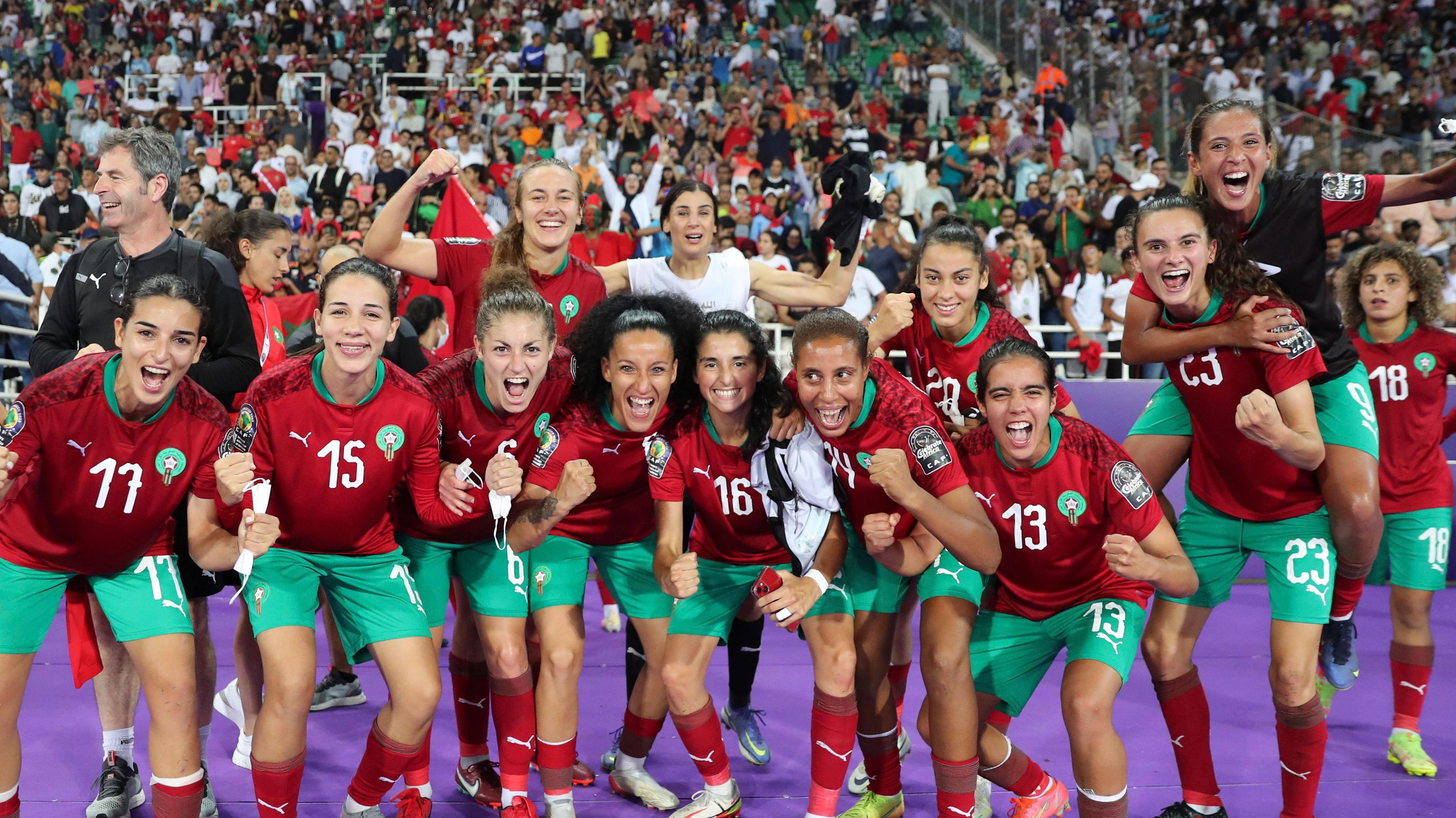 A group of Morocco players stand with their backs to fans in a stand and celebrate winning a match at the 2022 Women's Africa Cup of Nations by looking into the camera, clenching fists and shouting