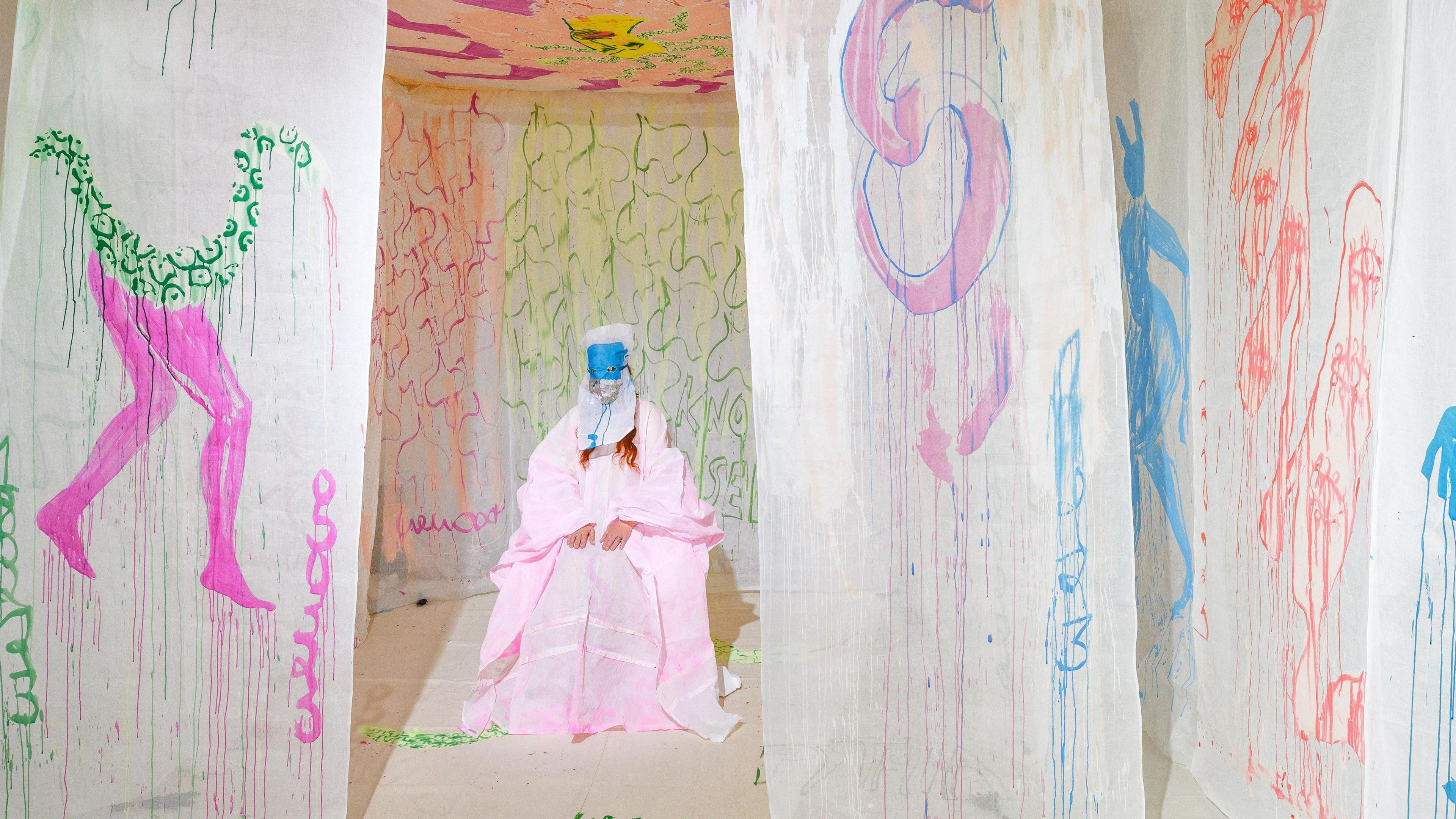 There are pieces of fabric hanging down around a central artwork of a seated figure. The fabrics have colourful paint on them- one of them shows a green snake with pink human legs, inside the circle of of hanging fabrics the figure is dressed in a pale pink dress and shawl, with a white and blue mask covering the head.