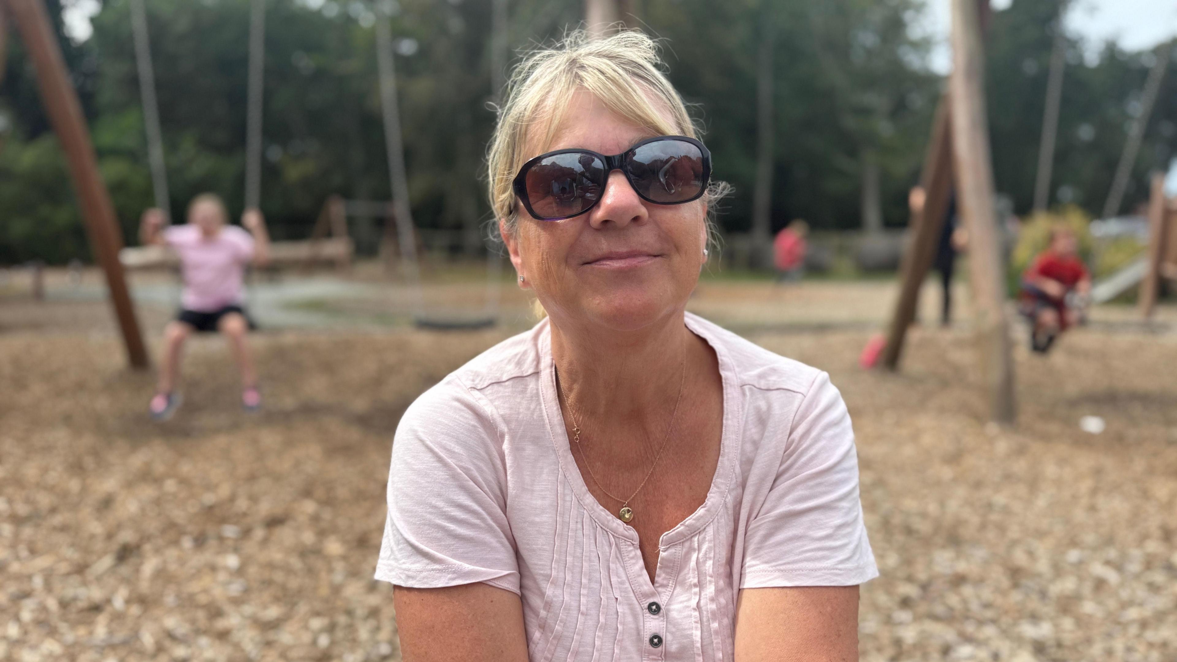 a woman in her late 50's wearing a light pink cotton top. She wears round sunglasses and has blonde hair up in a ponytail