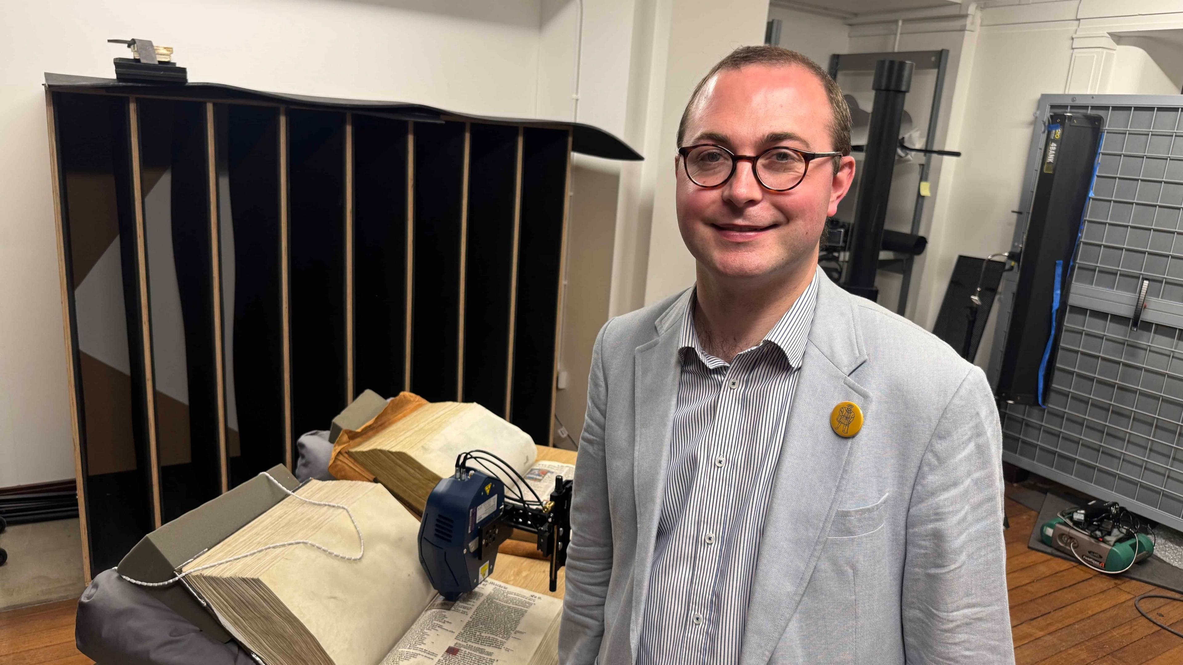A man in a grey suit with glasses stands in frount of technology which is scanning ancient Bibles.