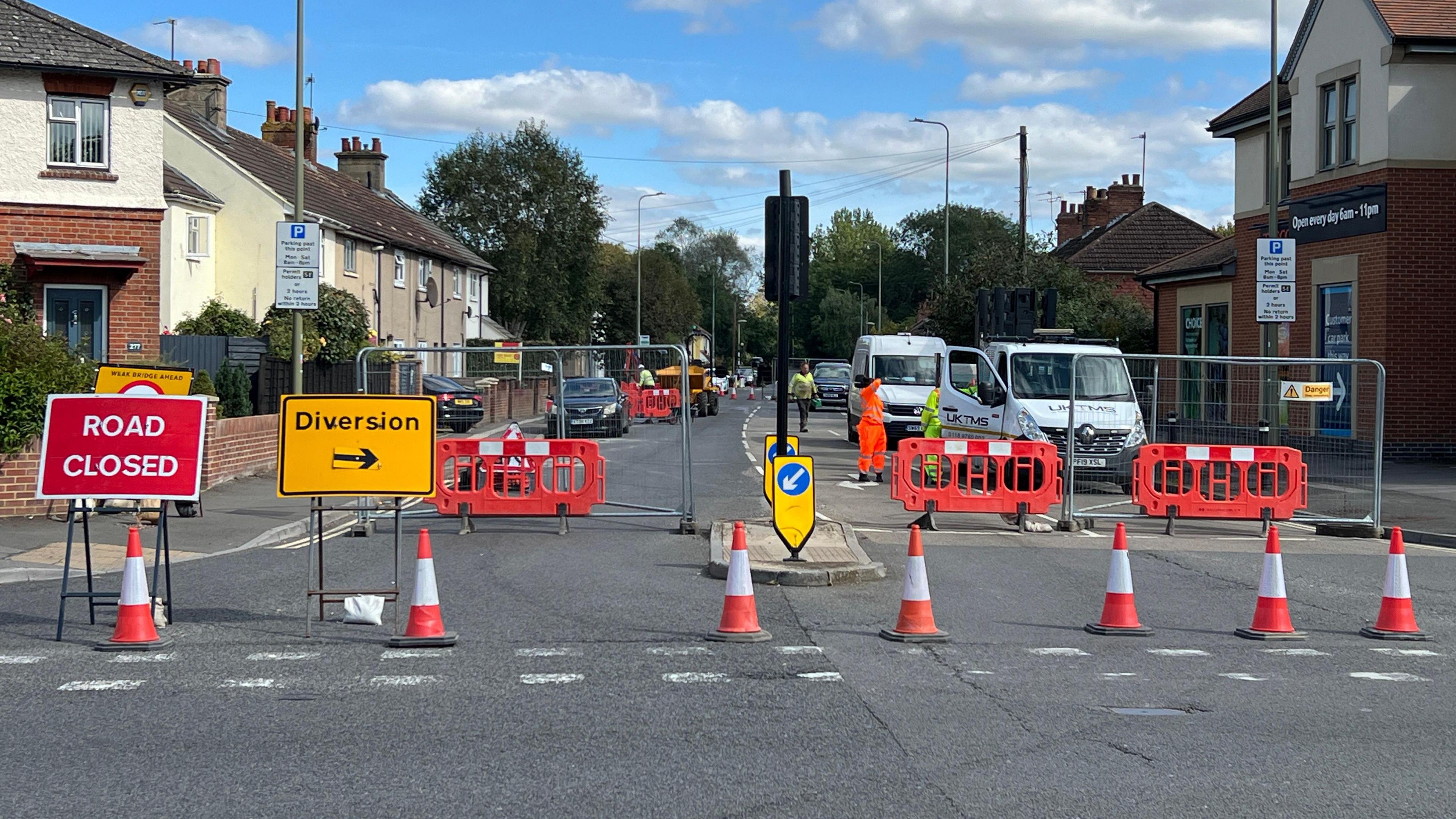 Signs reading Road Closed and Diversion alongside traffic cones in front of a closed road. A few workers in hi vis clothing and a few vehicles can be seen behind the barriers.