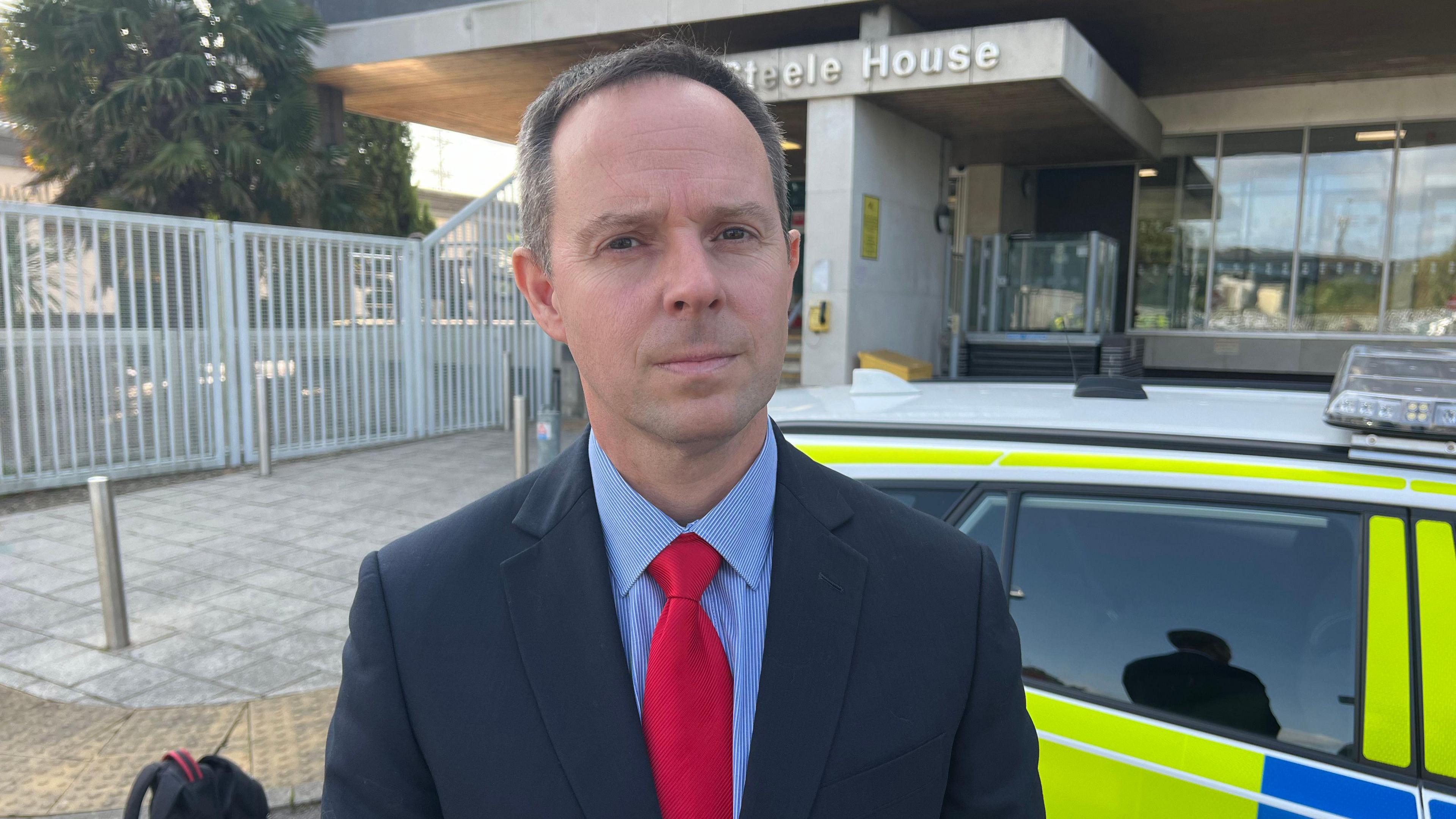 A man wearing a suit, blue shirt and red tie, stood in front of a police car, staring at the camera