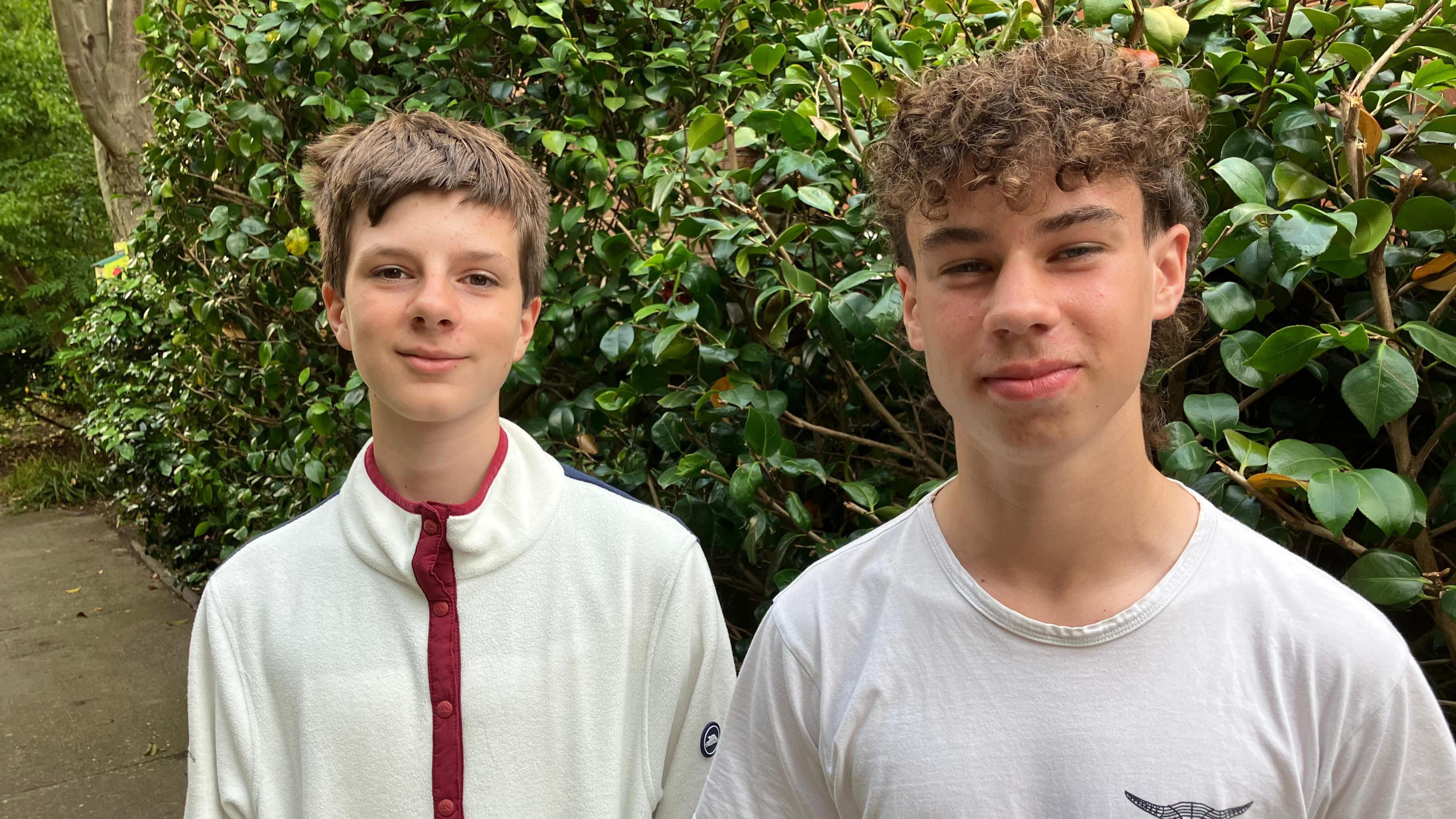 Two teenage boys smile at the camera, standing in front of a hedge. A tree is also in the background.