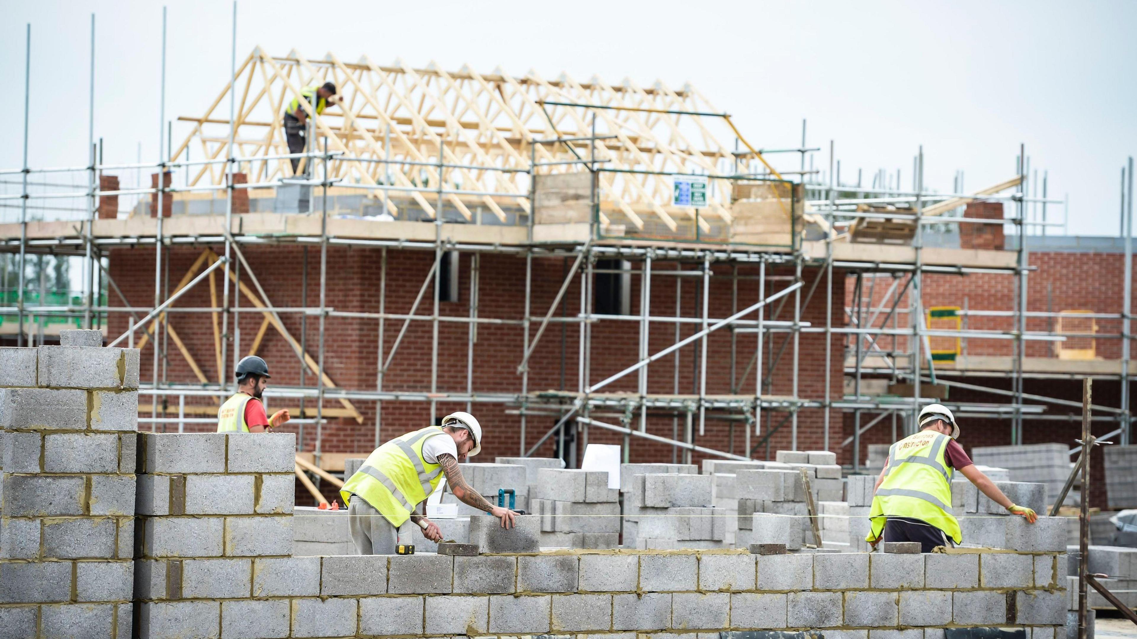 Several labourers lay a breeze block wall with an under-construction house with roof timbers and scaffolding behind them.