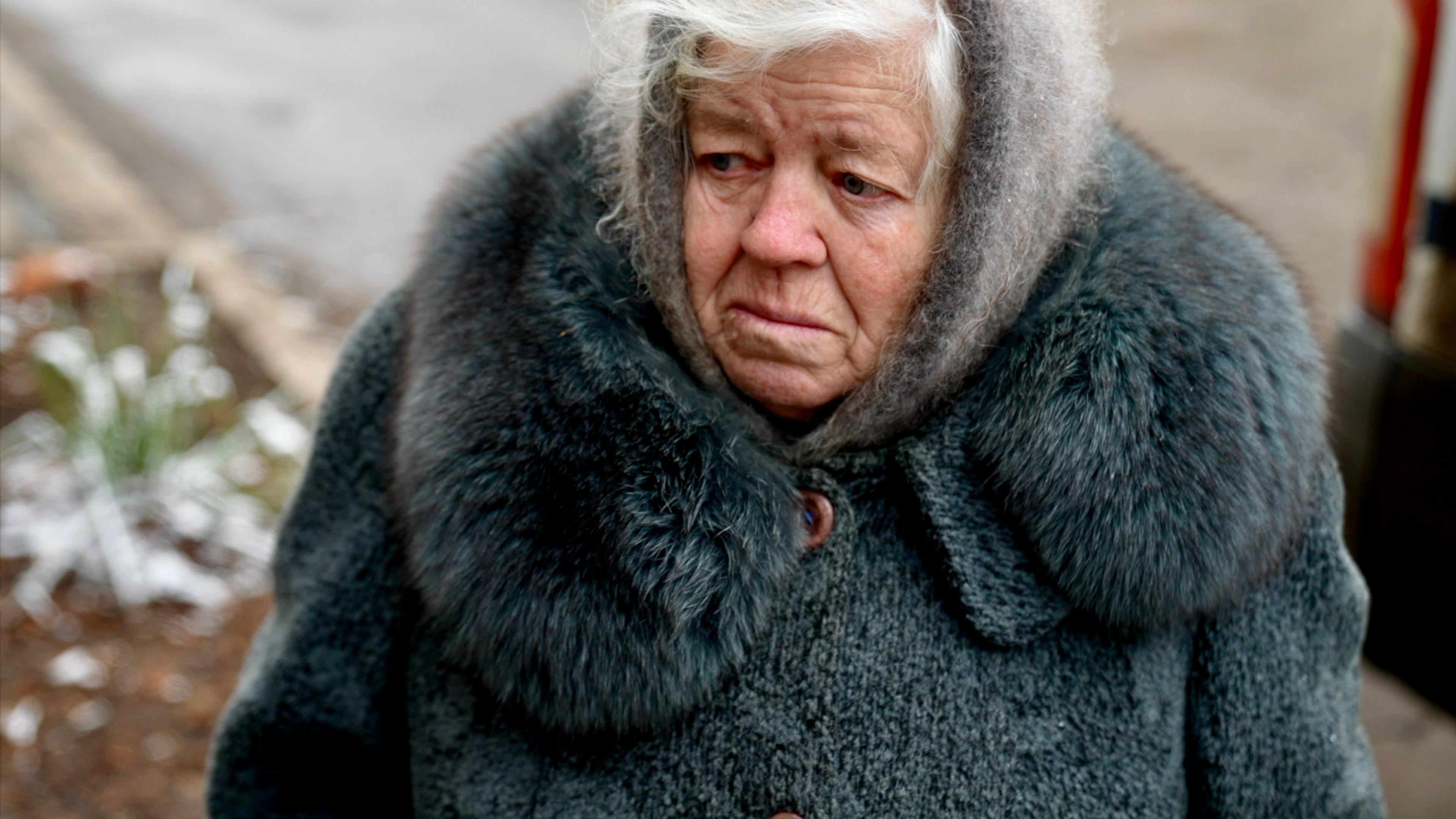 Lyuba stands outside wrapped up in a grey furry coat and hood with a sad expression on her face