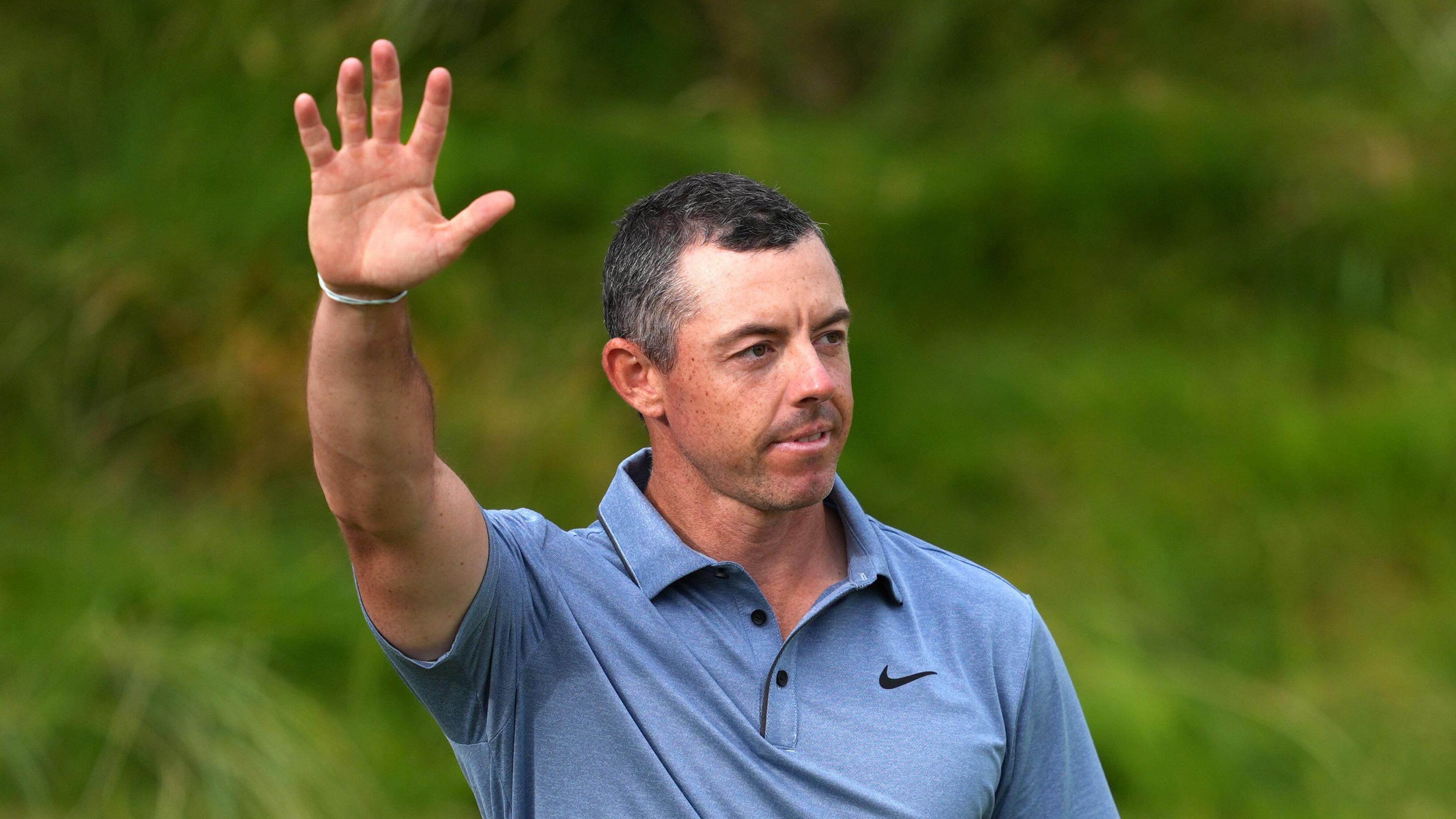 Rory McIlroy, with short black hair, wearing a light blue Nike golf shirt. He is holding his hand in the air.