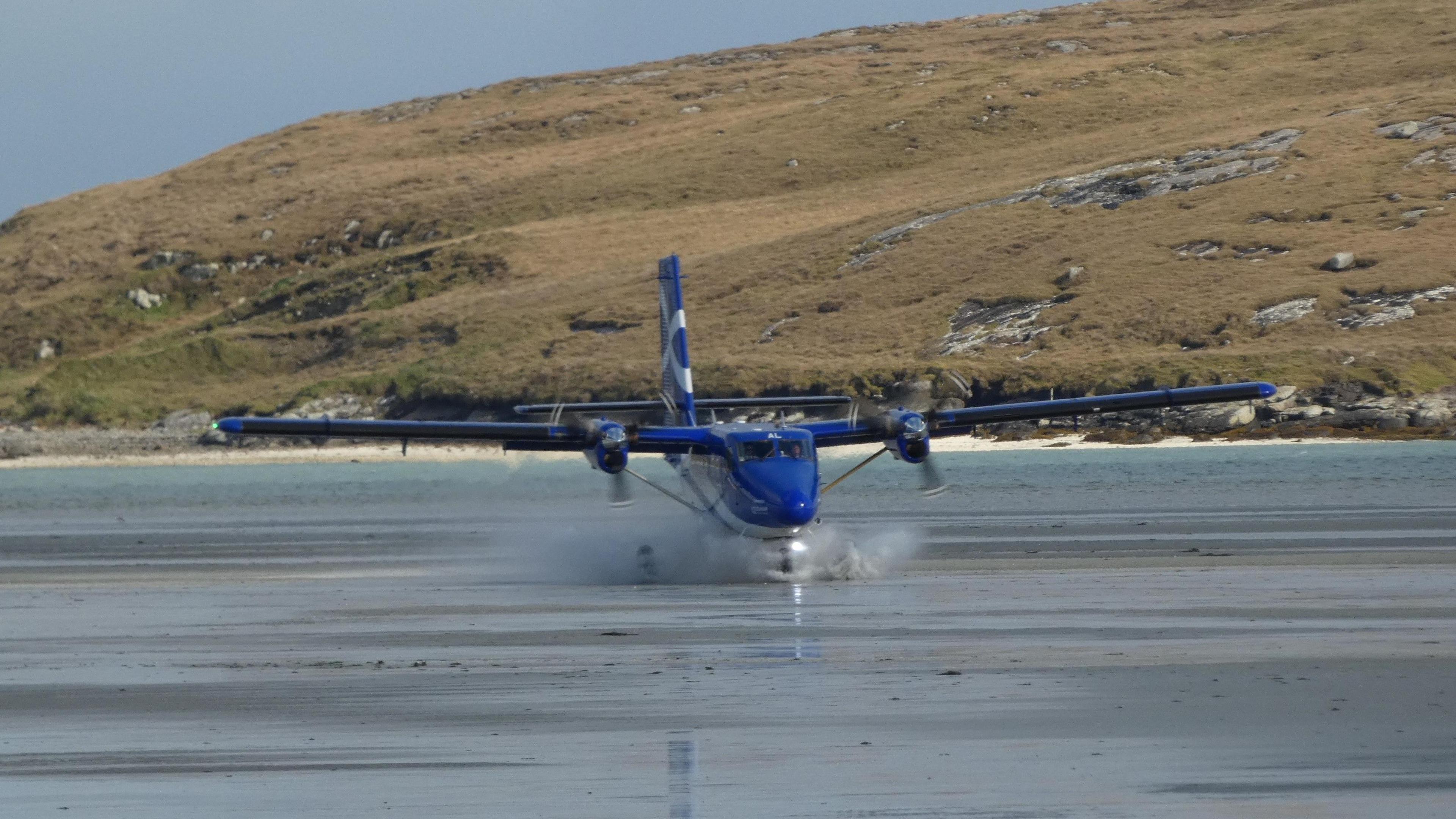 A blue Loganair Twin Otter small plane lands in the shallow water on the beach in Barra. The water splashes around the landing gear and the beach is behind the aircraft.