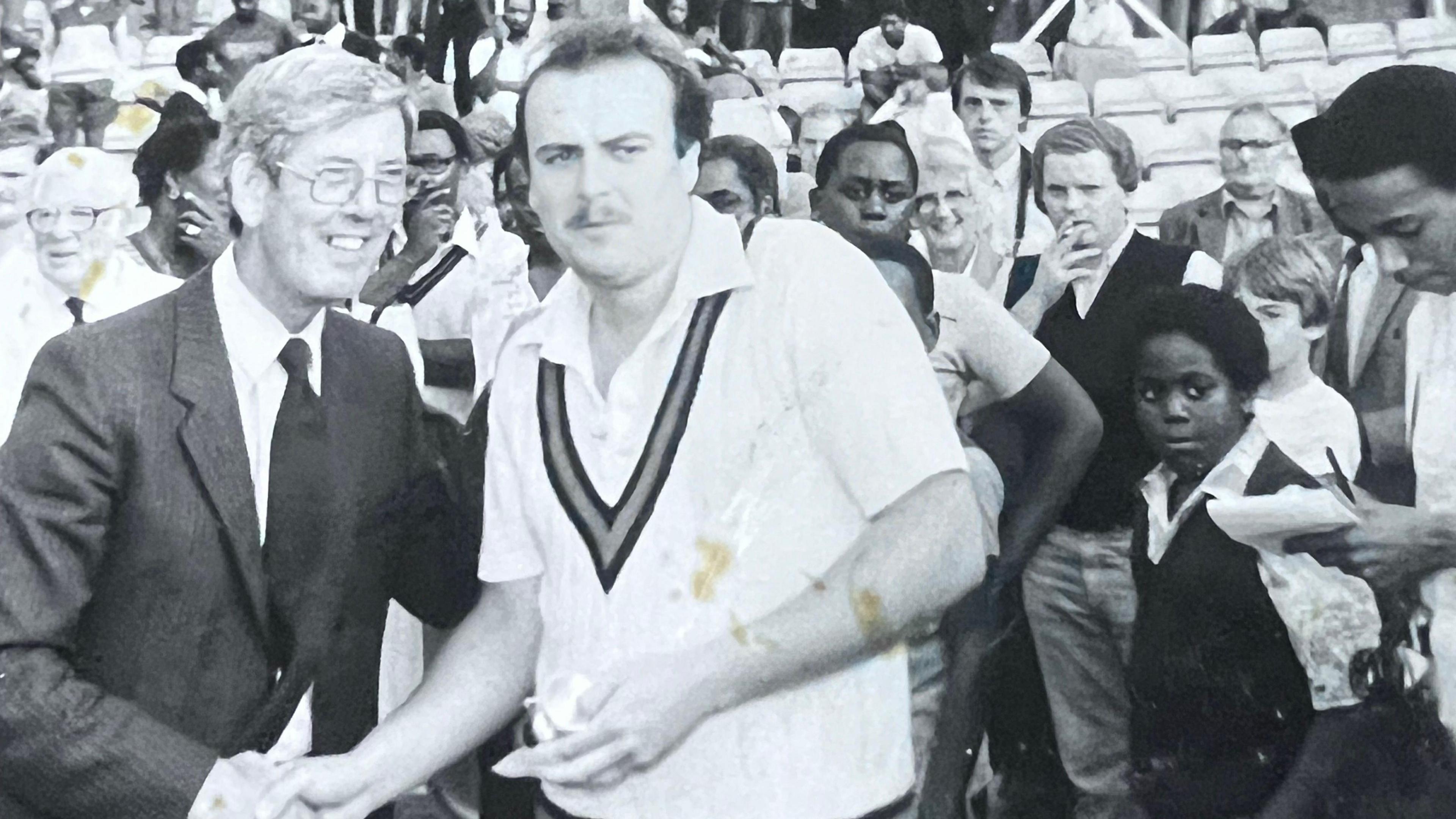 A black and white image from 1982, showing A Met police officer, taking part as a cricket player, shaking hands with another man as spectators, including men, women and children, stand watching behind them. The man in the centre of the front row holds a prize shield. The photograph is taken at a cricket ground.