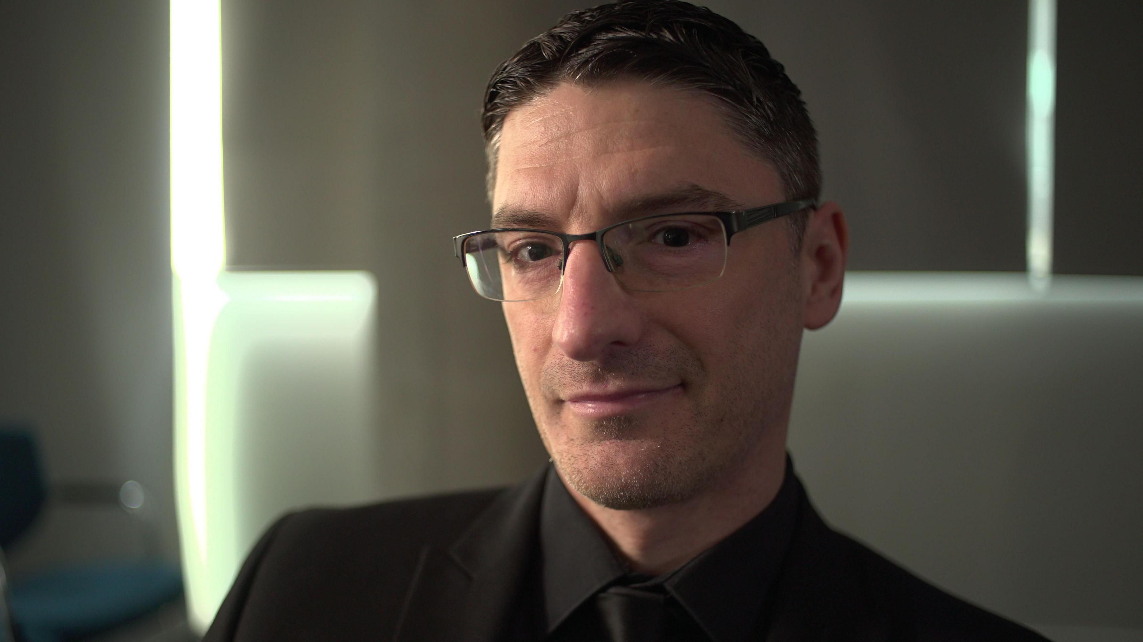 Mark Collett, who set up the far-right Patriotic Alternative, is pictured looking into the camera with a neutral expression, wearing glasses, a black shirt, black tie and black blazer.