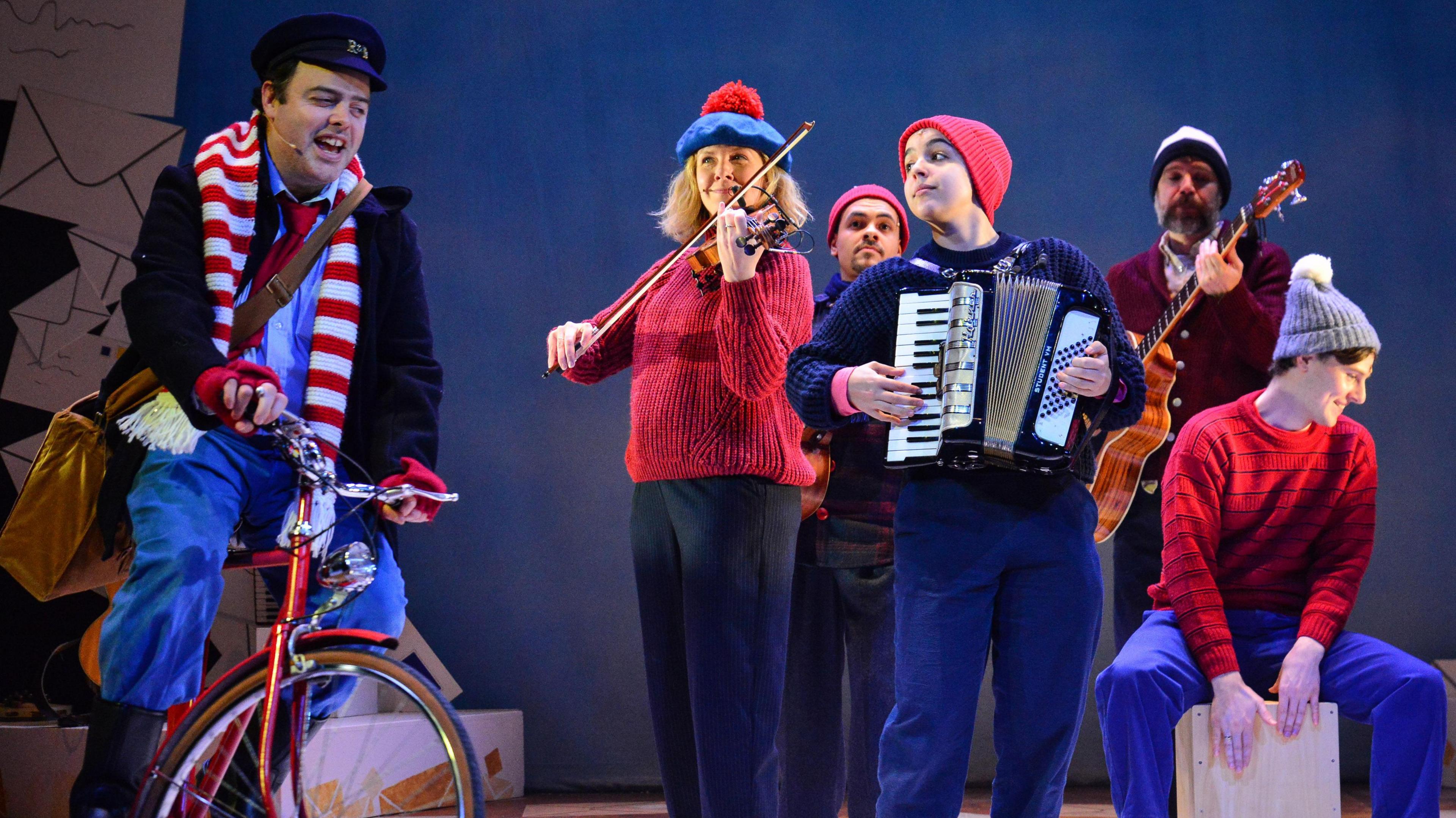 A man with a scarf and a hat rides a bike on stage a group of musicians play near him