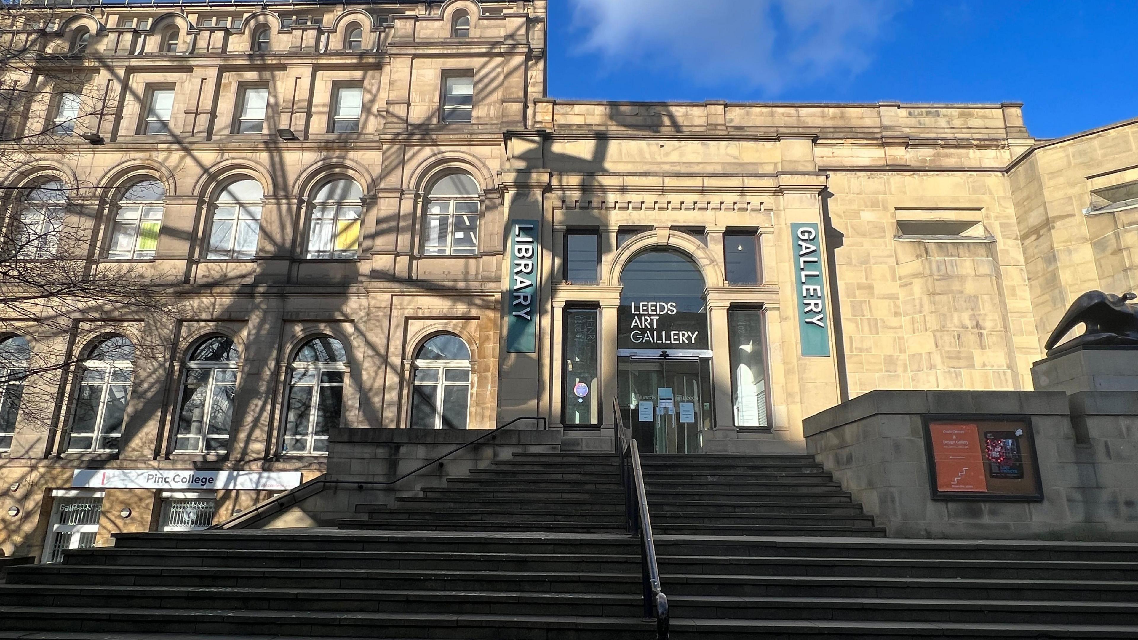 Exterior view of Leeds Art Gallery, featuring a stone façade with arched windows, a wide staircase leading to the entrance, and vertical banners labelled "LIBRARY" and "GALLERY".