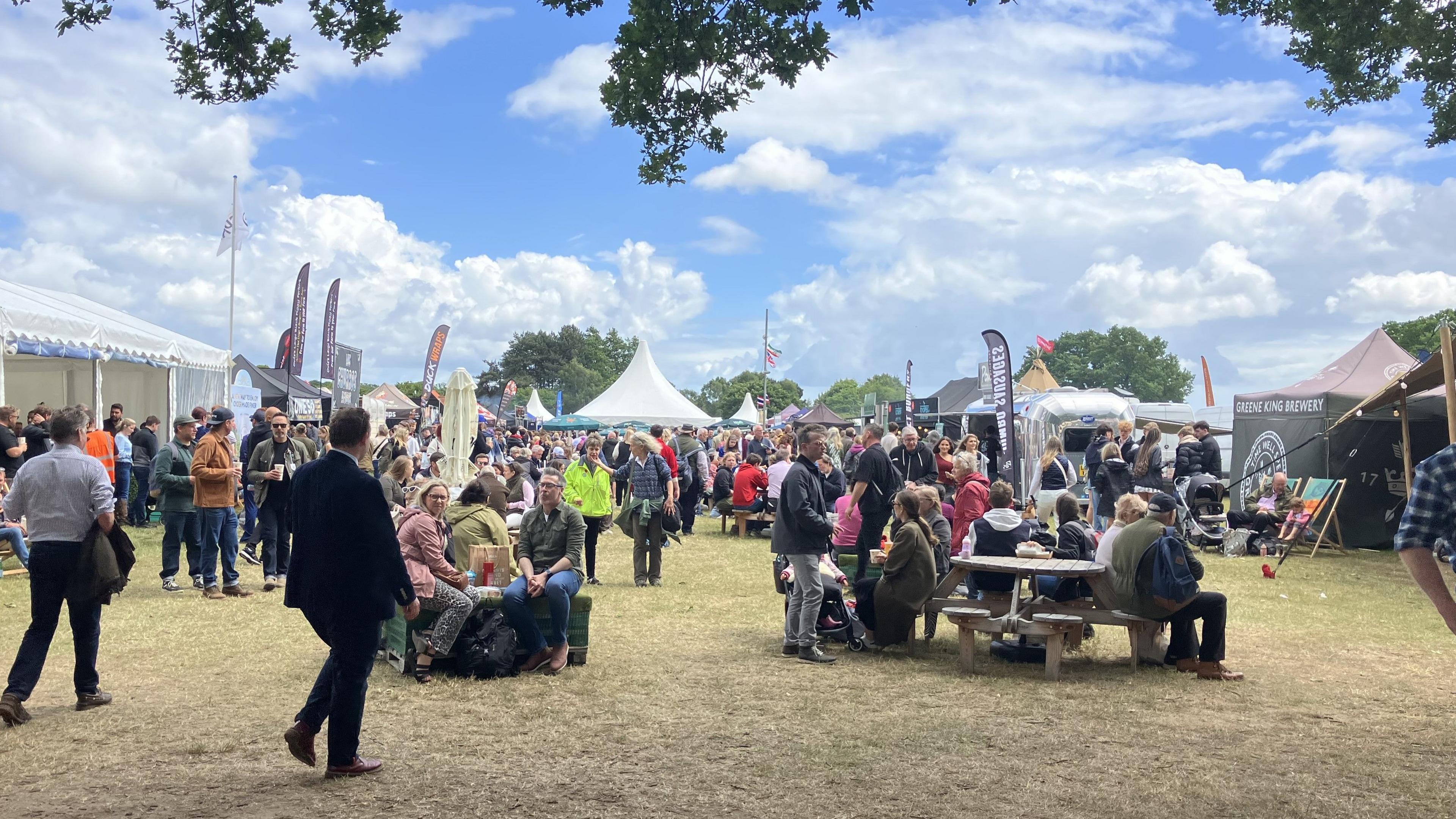 Crowds of people within a open space at the Suffolk Show. Some people sit on benches and talk while other meander around them. 