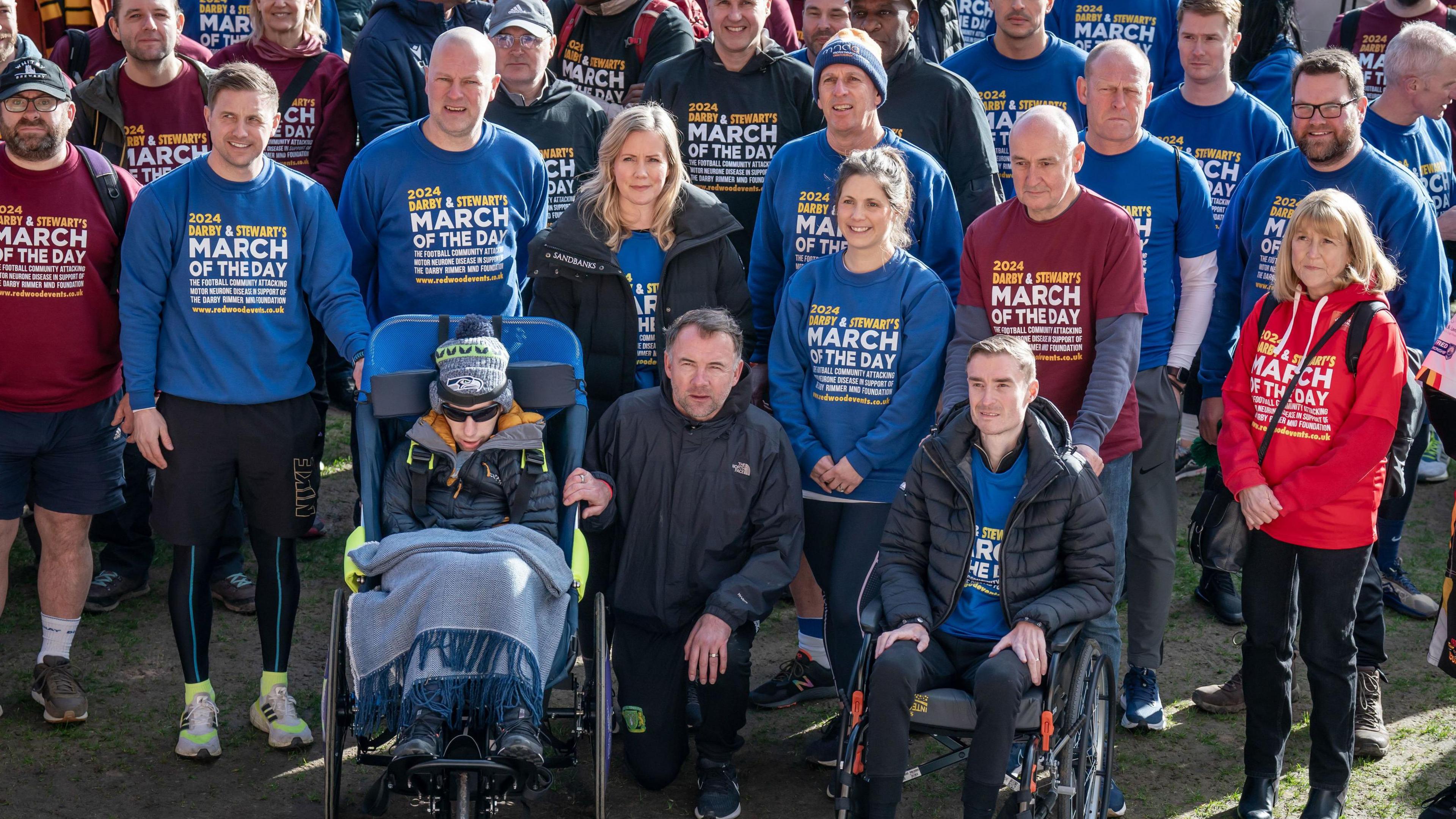 A large group of people gather wearing branded "march of the day" sweaters. Marcus Stewart crouches front and centre dressed in a black tracksuit between Rob Burrow and Stephen Darby.