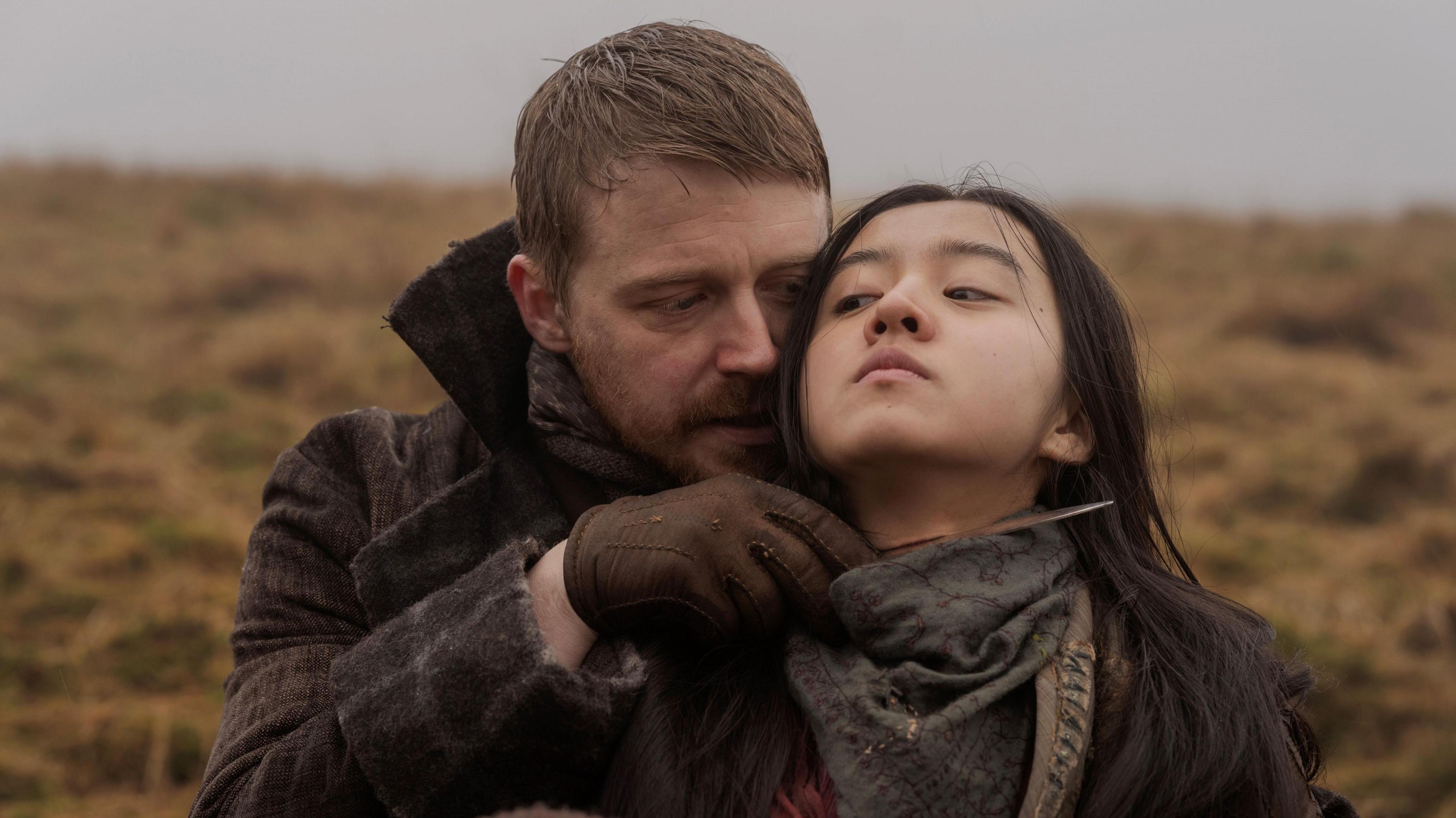 Still from upcoming film Tornado showing Little Sugar (Jack Lowden) and Tornado (Kōki) - Little Sugar holds a blade at Tornados neck as he stands behind her on a grass covered hill with grey skies behind them.