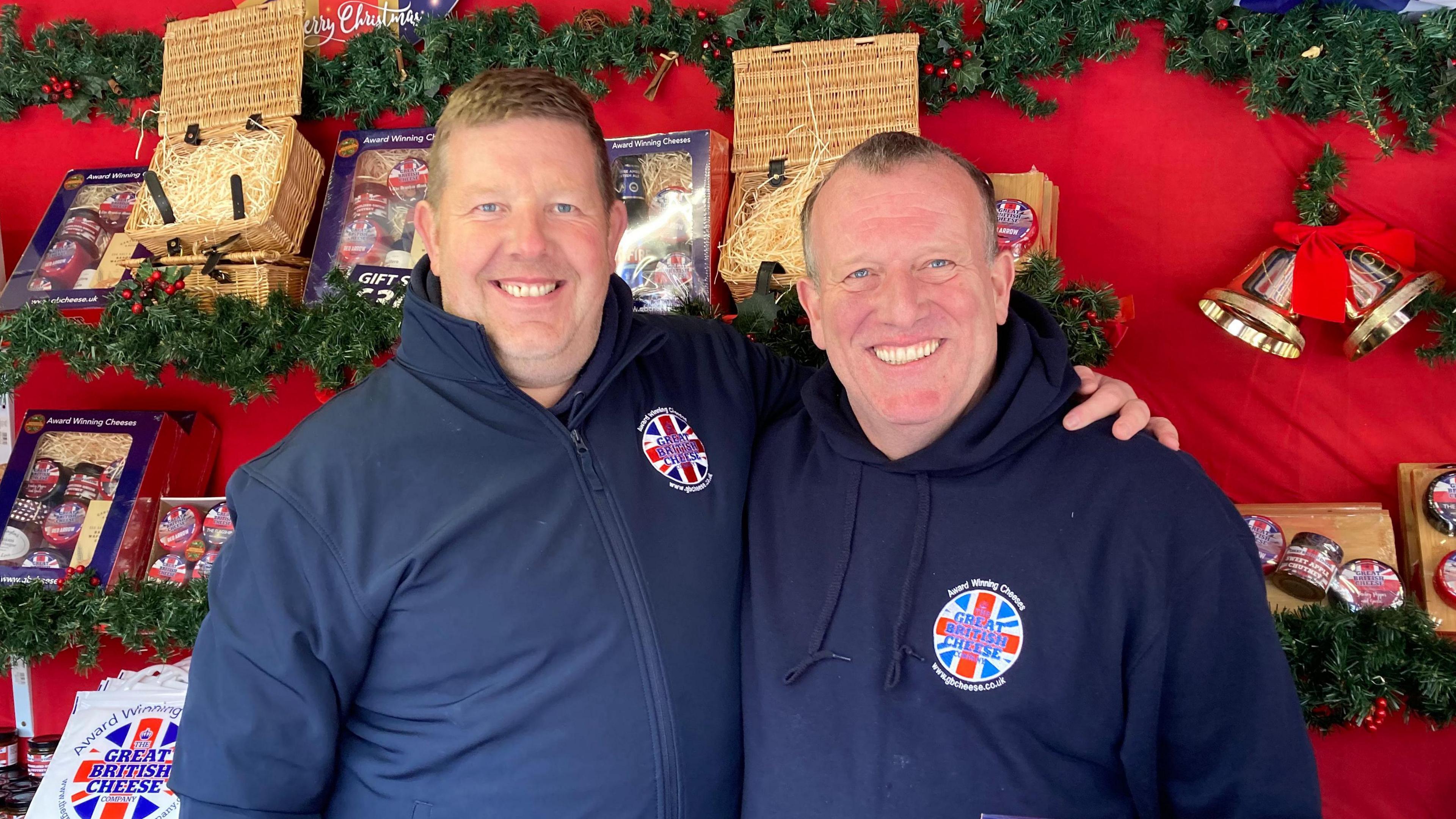 Two men stand in front of a cheese stall