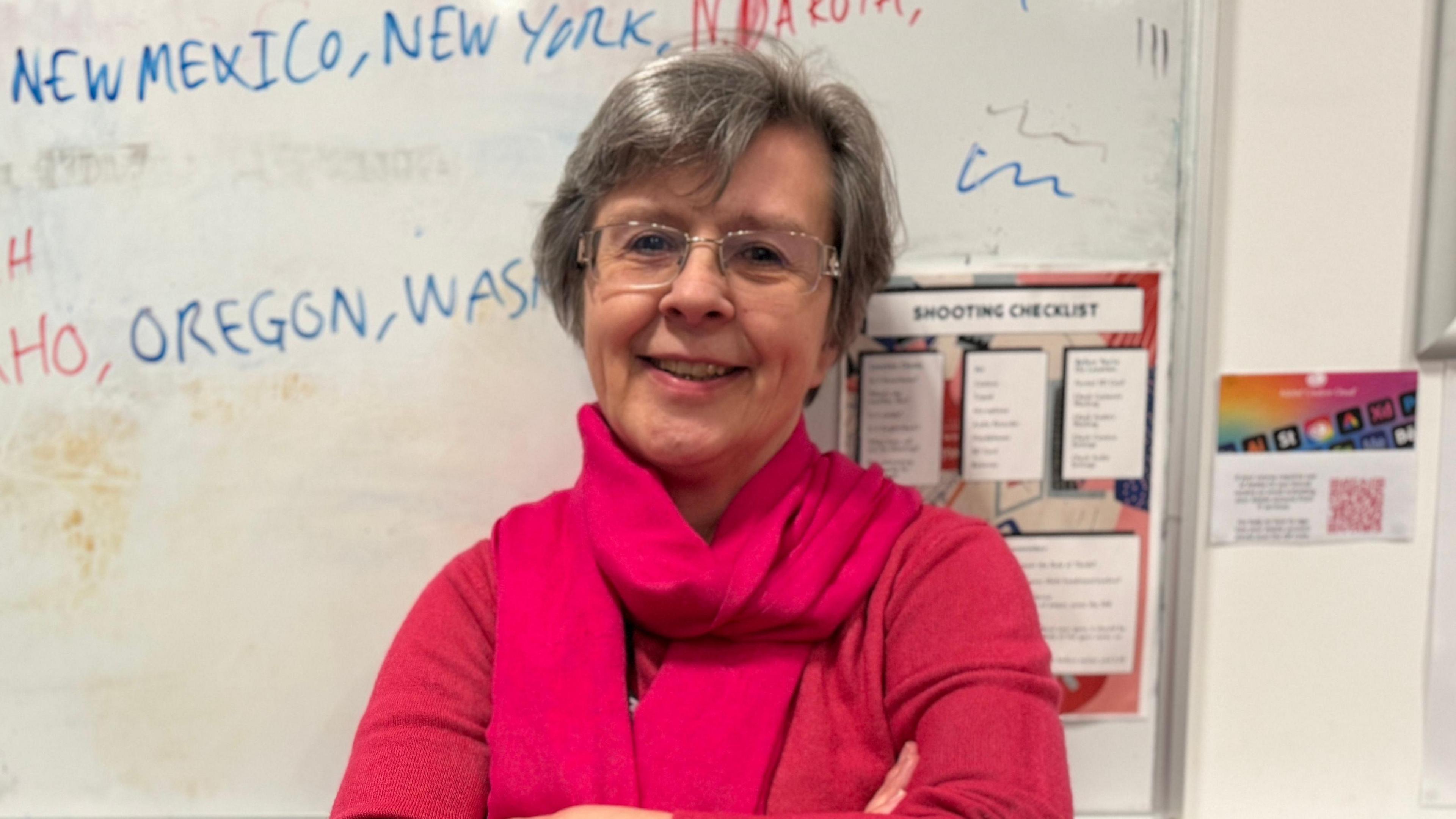 A woman smiling and looking directly at the camera, wearing a pink jumper and scarf with clear-framed glasses. 