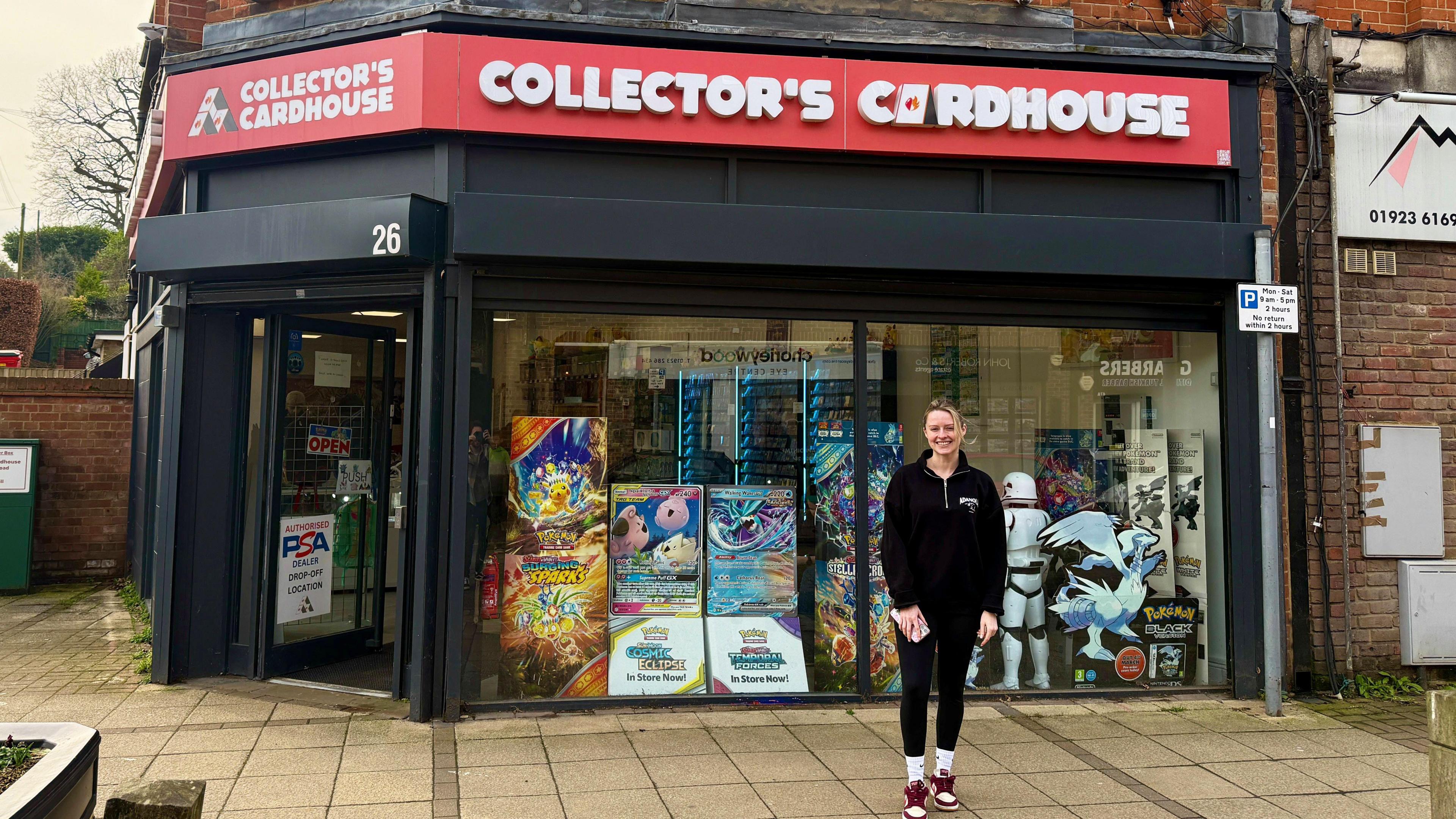 A young blonde woman is smiling outside a shop which has a large red sign saying "Collector's Cardhouse' above it