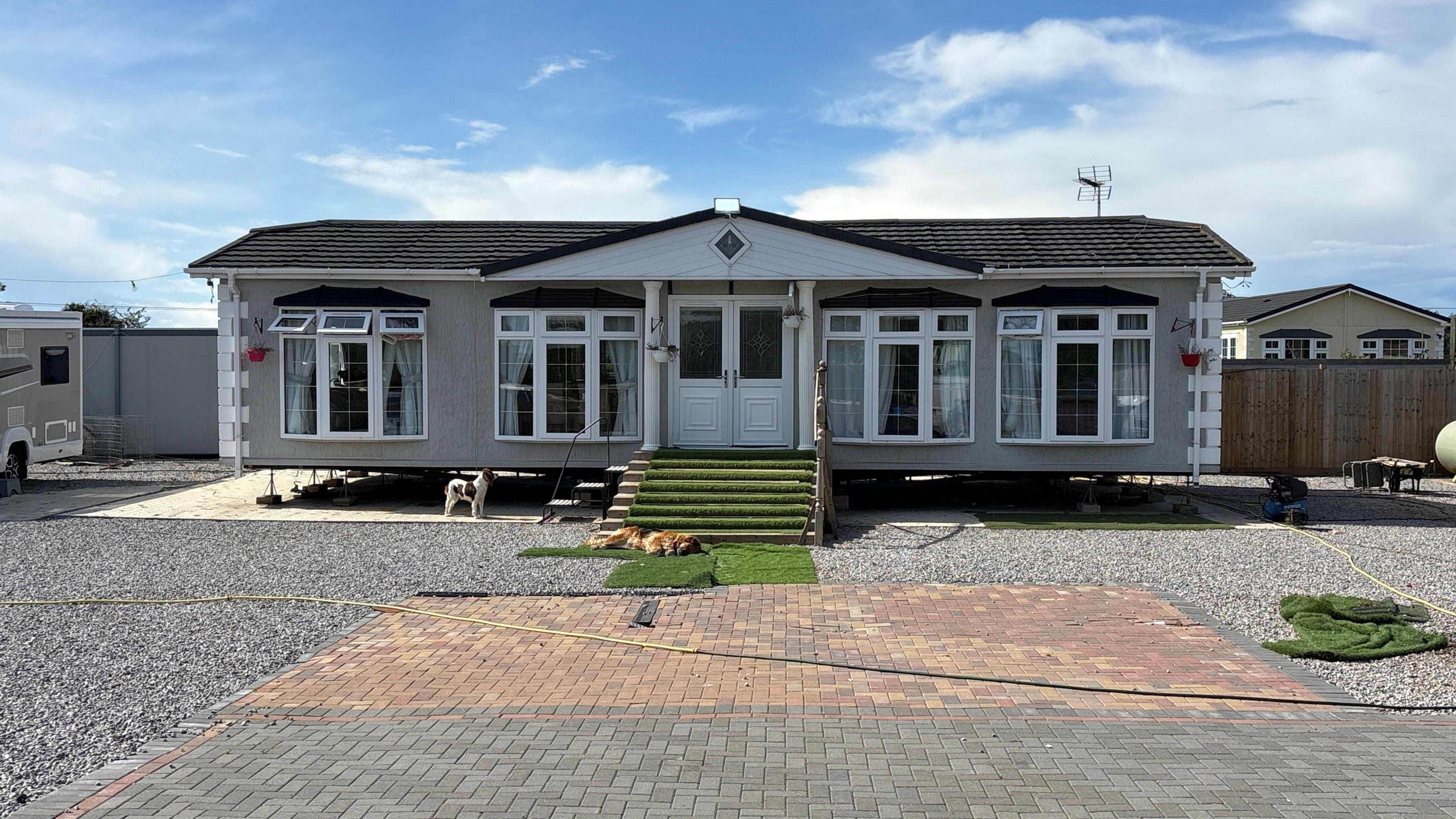 A wide shot of a static caravan. It is set back from where the camera is stationed, with an expanse of block paving and a stone driveway in the foreground. A small stairway leads up to the caravan's double, uPVC-type doors. Either side of the doors are two sets of windows, meaning across the front there are four sets of windows in total. The caravan is symmetrical and imposing against a light blue, partly cloudy sky. 