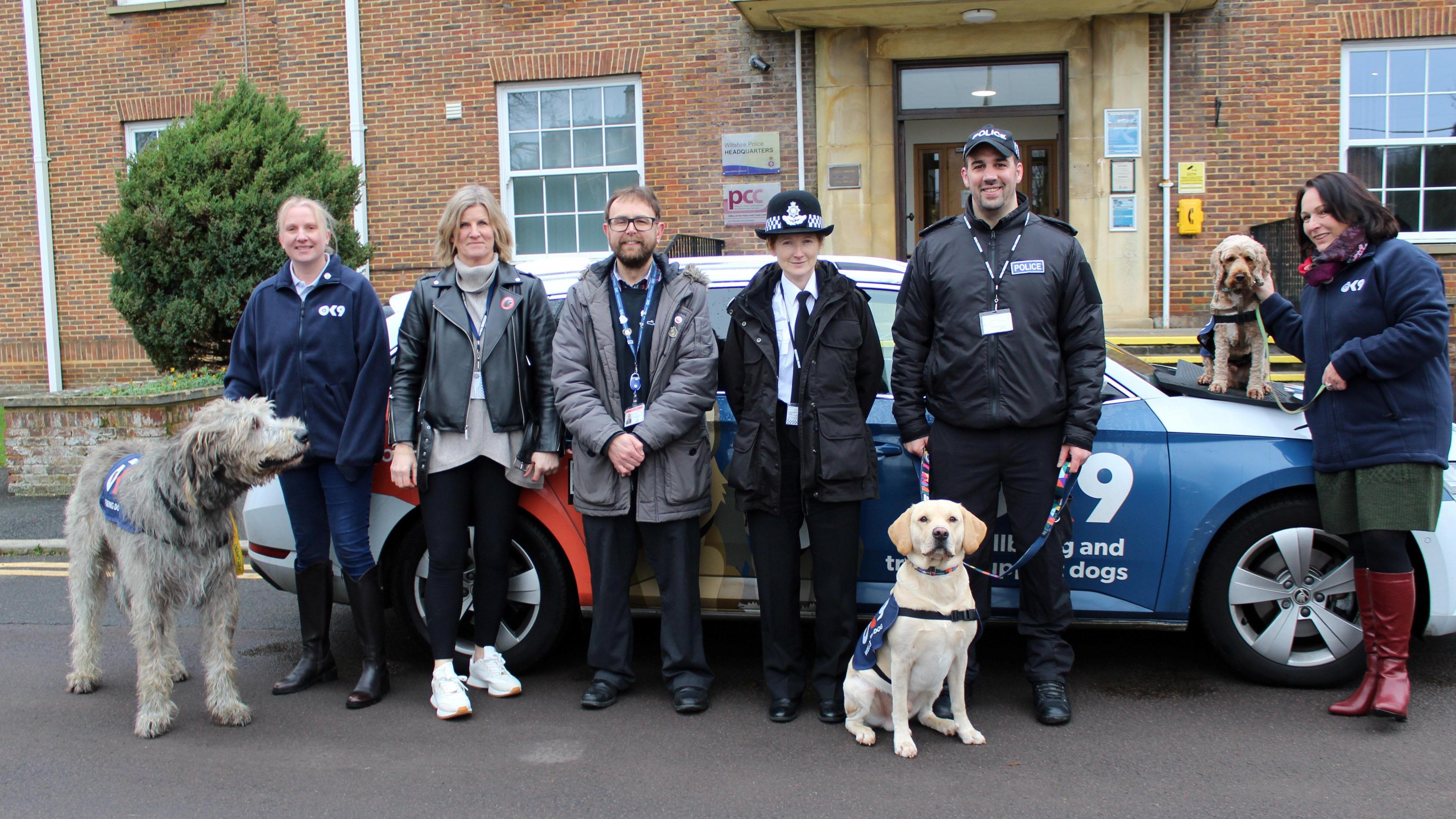 Six people stood outside a police station. There's dogs sat like good dogs in front of the police car.