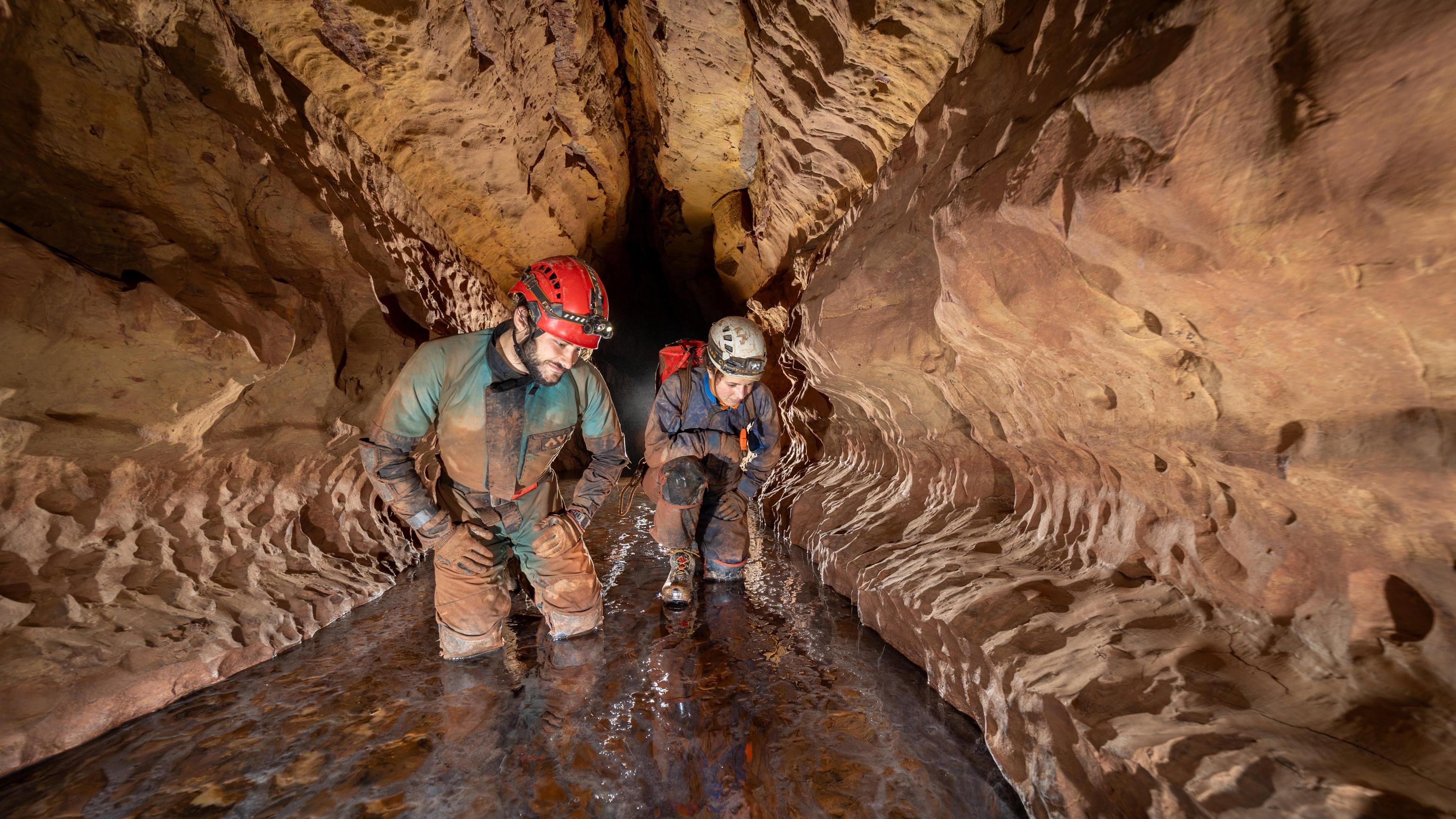 Two cavers from the exploratory team studying the rock formations and smiling. They are both wearing helmets and waterproofs standing in water up to their knees.