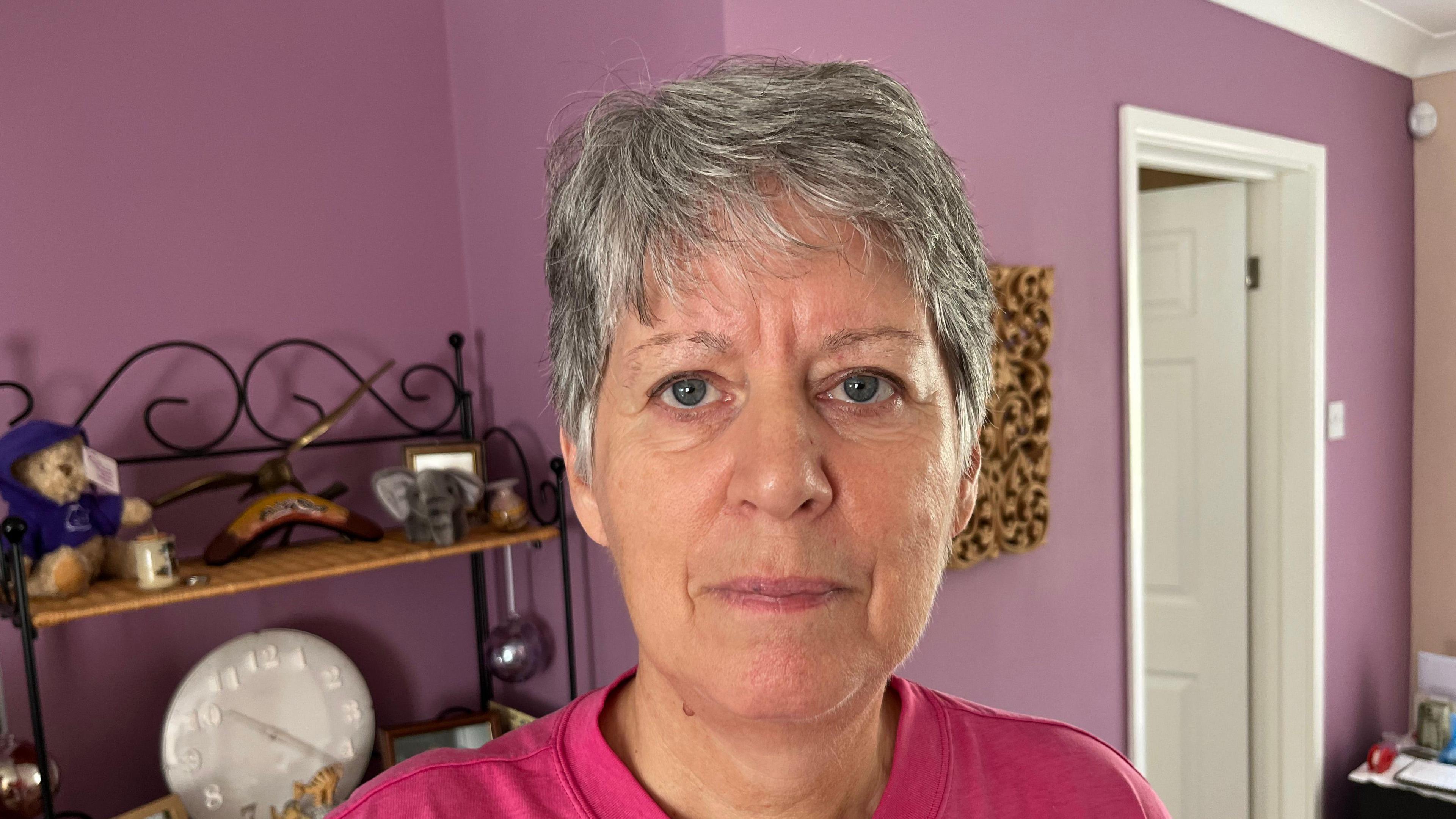 Mandy Amory has short grey hair with a pink round collared T-shirt she is standing in a living room with dark pink walls and a shelf with various ornaments on.