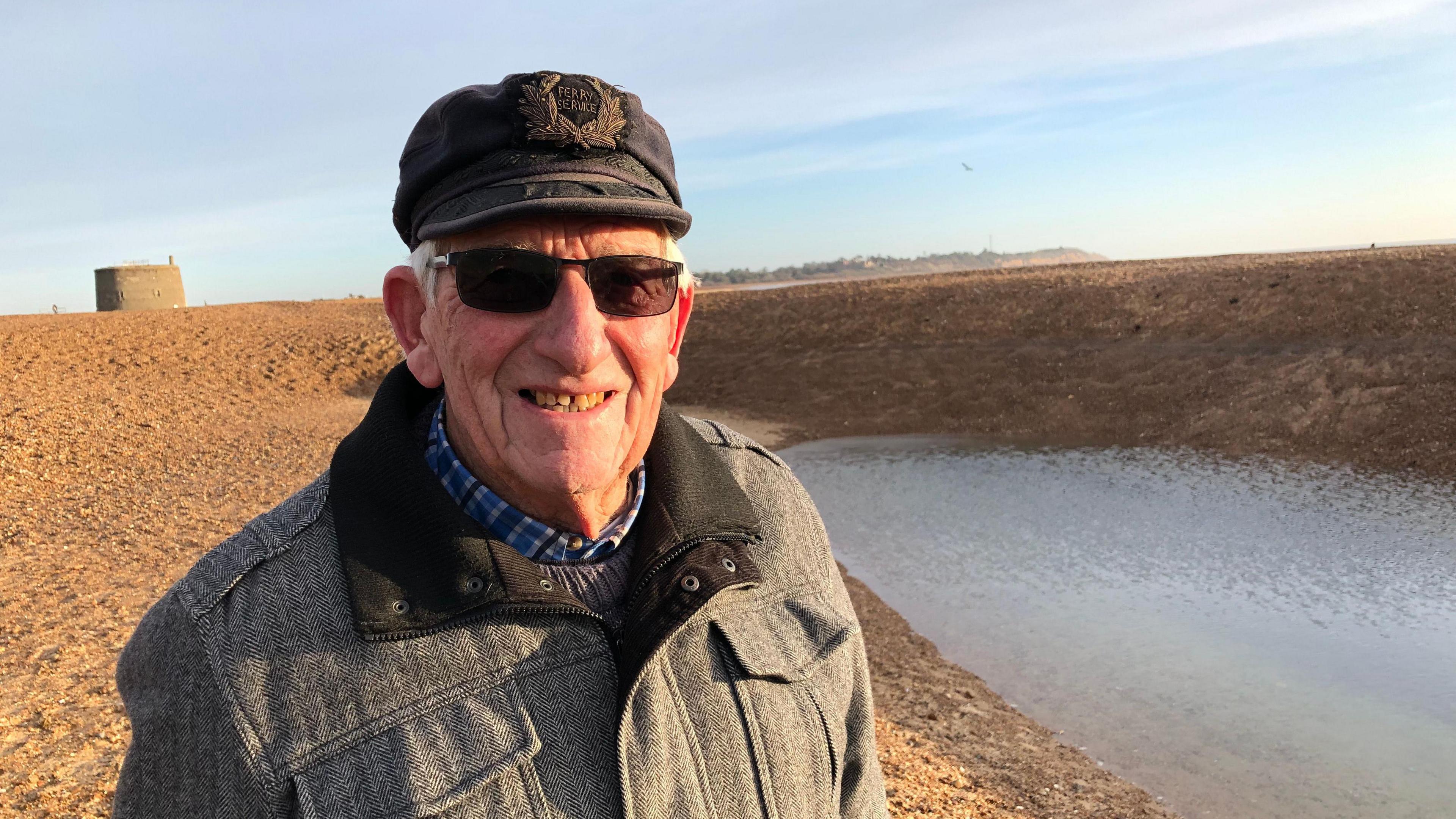 John White smiles at the camera in front of the hole that has appeared on Felixstowe beach. He wears a cap with sunglasses, and a grey coloured coat.