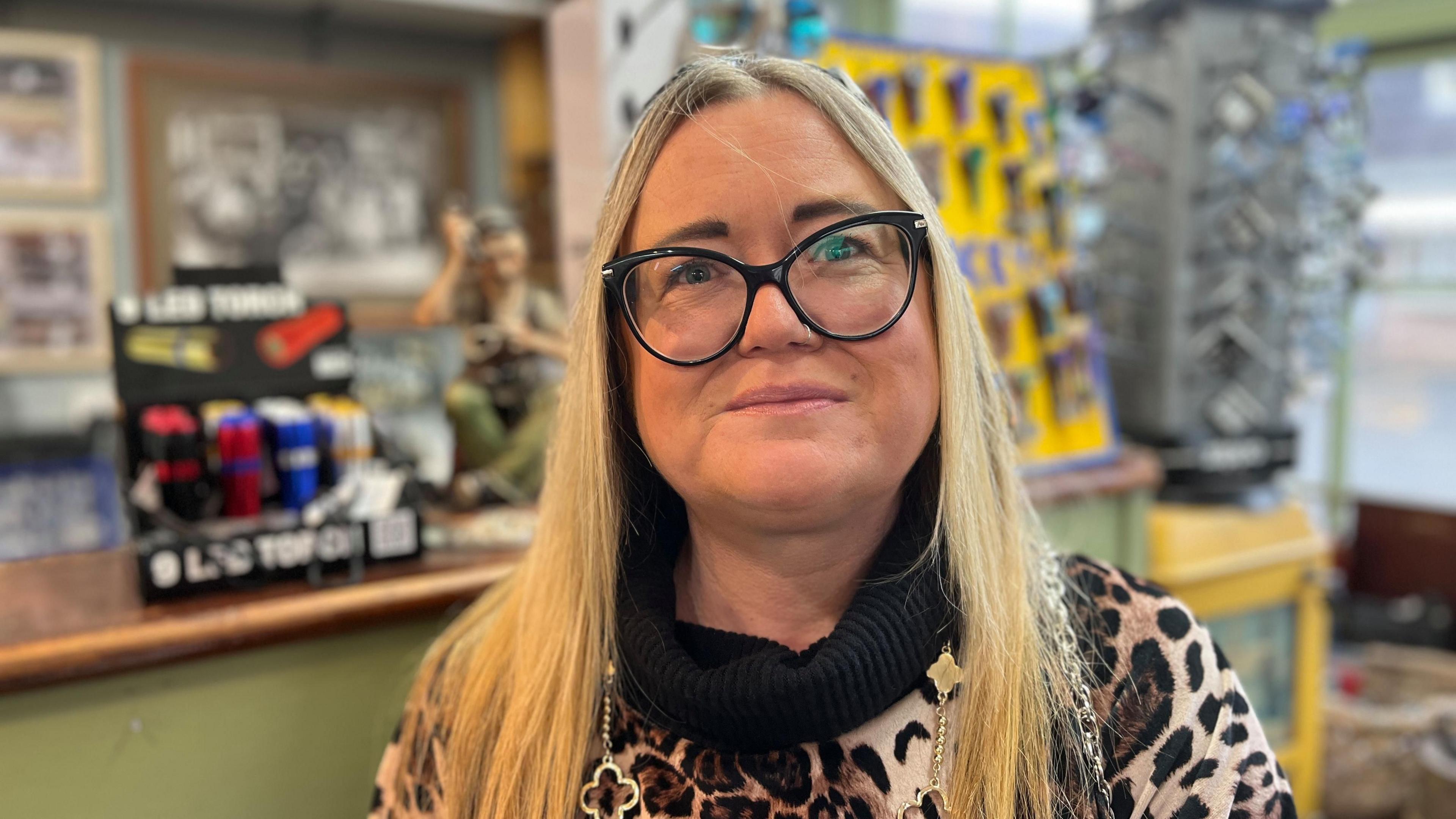 A photograph of a middle-aged woman, smiling at the camera. She is white with long blonde hair and wearing thick-rimmed glasses with a leopard print jumper. She stands in a shop, with indistinct items for sale in the background.