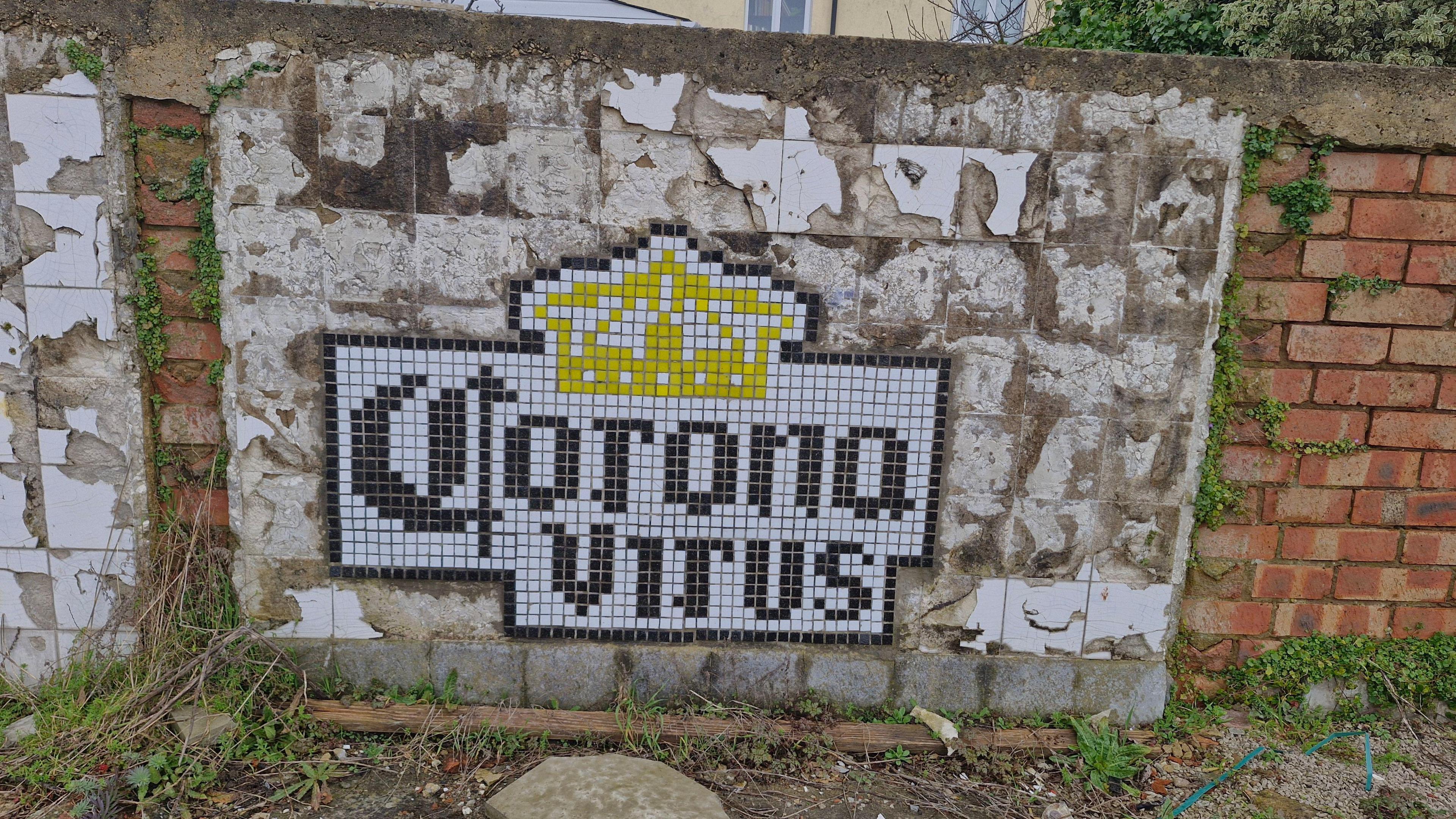 An artwork on a wall with the words Corona Virus and a golden crown in the style of tiling - weeds, crumbling plaster and red bricks also visible 