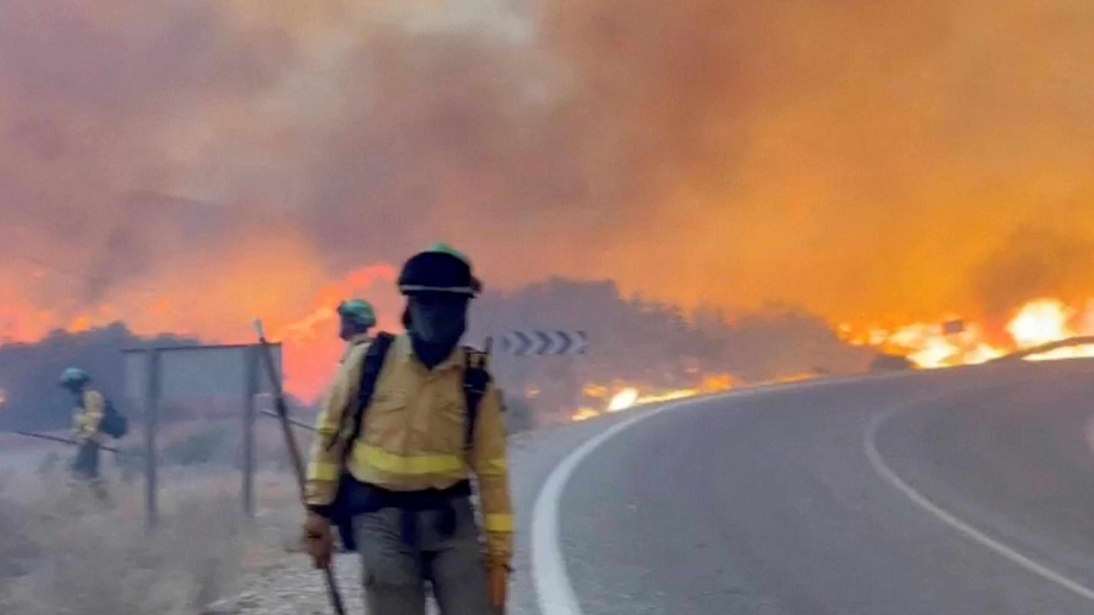 A masked firefighter stands next to a road in Spain while a wildfire rages behind them. 
