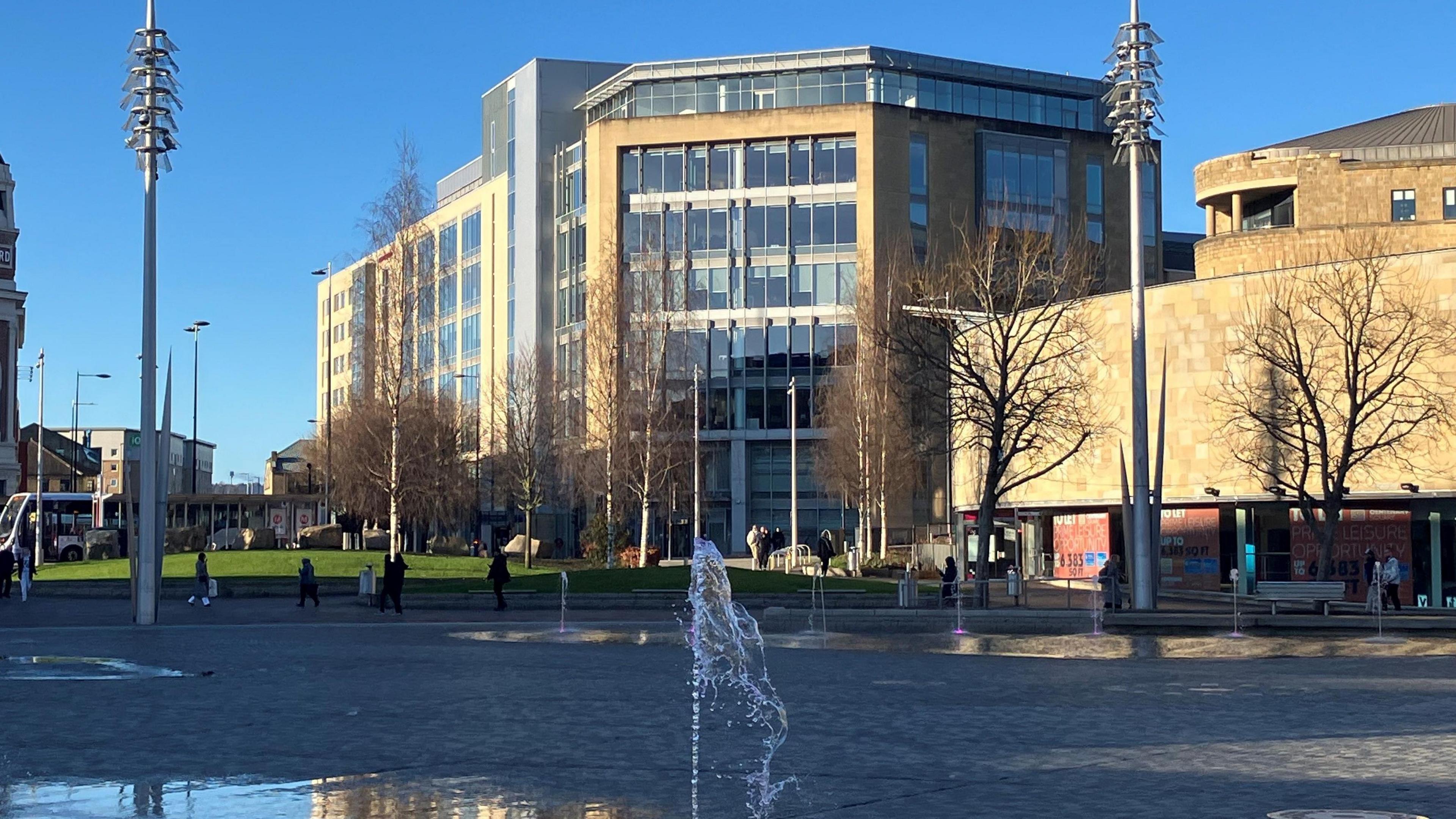 A modern city centre building pictured in sunshine from behind a fountain feature.