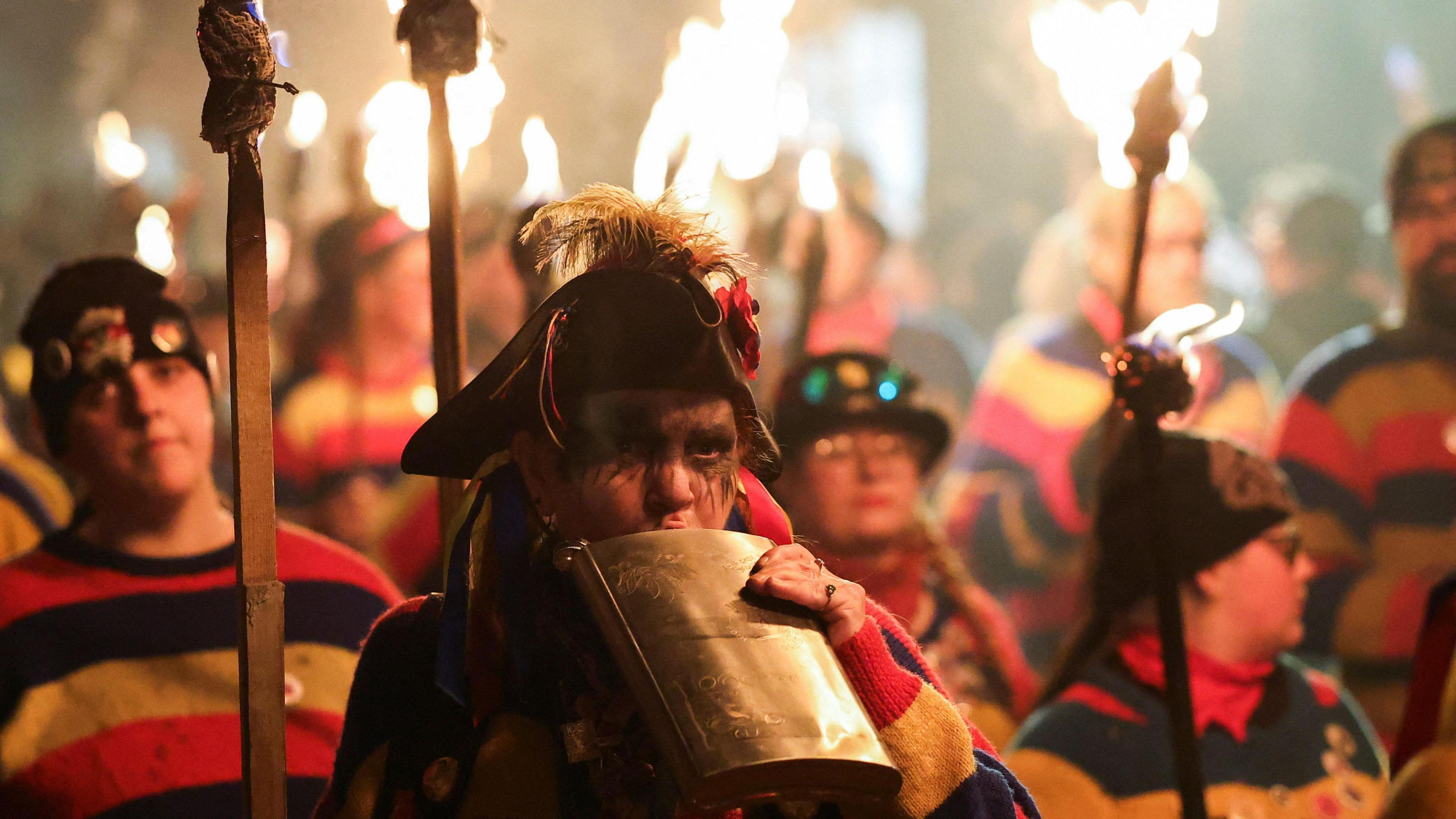 Many people in stripy costumes hold burning torches. One is drinking from a massive hip flask.
