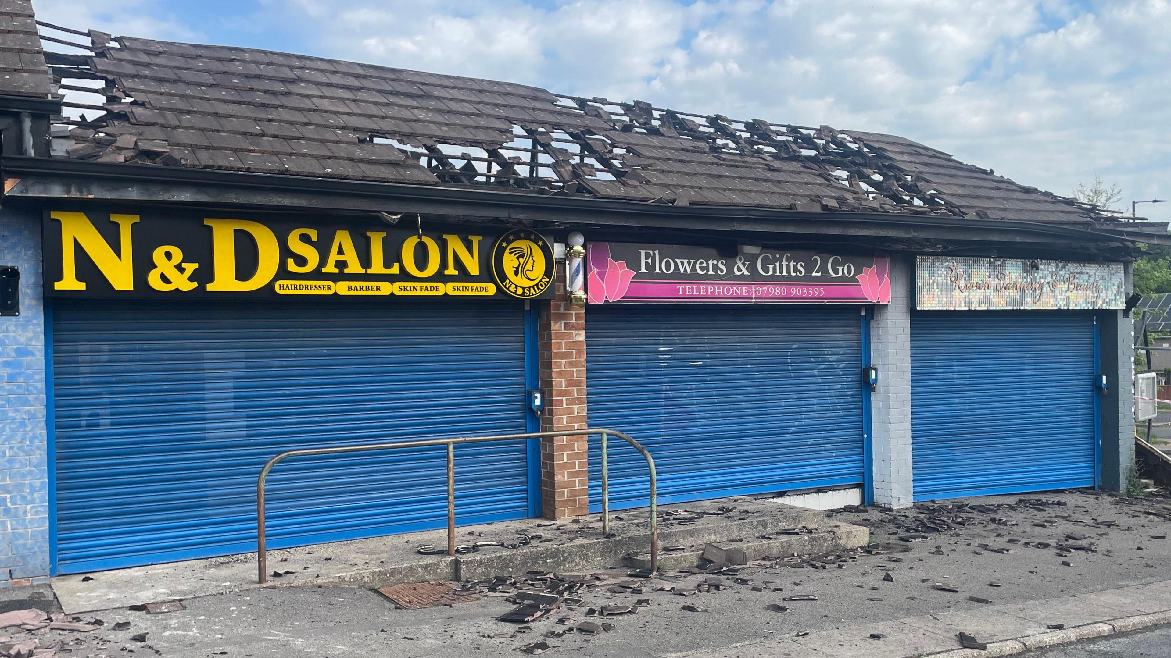 Front view of the shows, all with shutters down and the roof damaged. In places it has almost entirely fallen through.